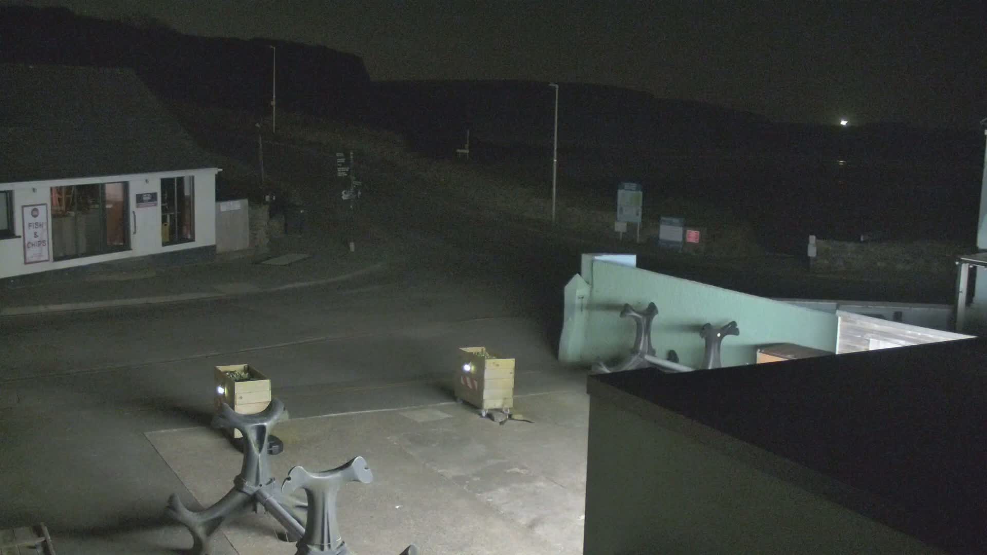 At night, under a mostly clear sky, a small town square is sparsely lit, showing two wooden crates and several dark metal bollards near a small building.