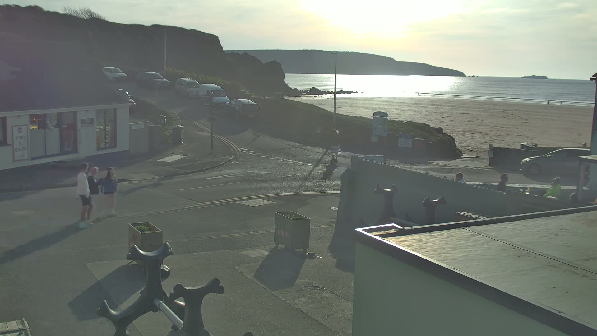 A seaside street scene at sunset shows a sandy beach, a calm ocean, parked cars, pedestrians, and a small building.