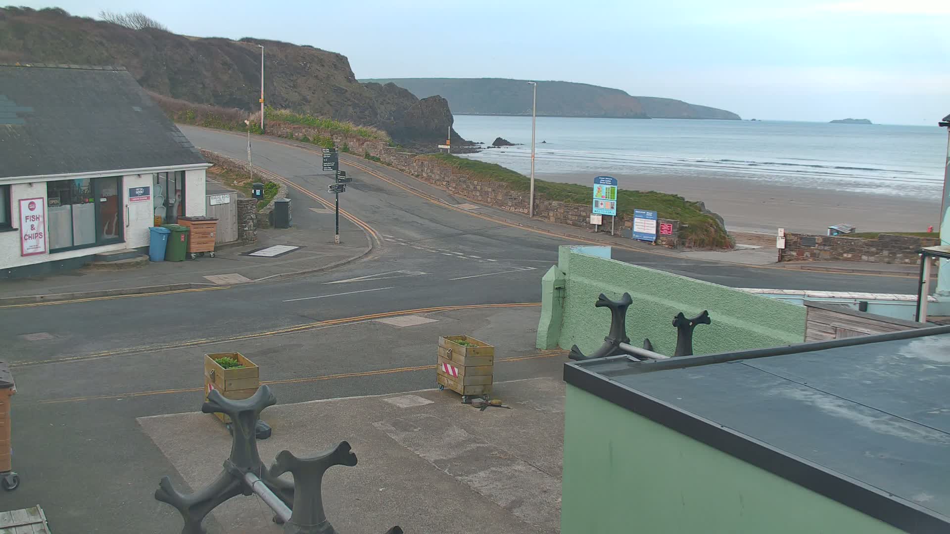 A coastal road curves toward a sandy beach with gentle waves under a cloudy sky, passing a small building with a fish and chips shop.