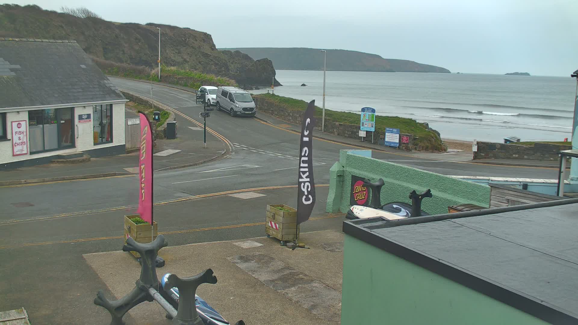 A coastal town scene on an overcast day shows a road intersection near a beach with small waves, a van, and buildings including a fish and chips shop.