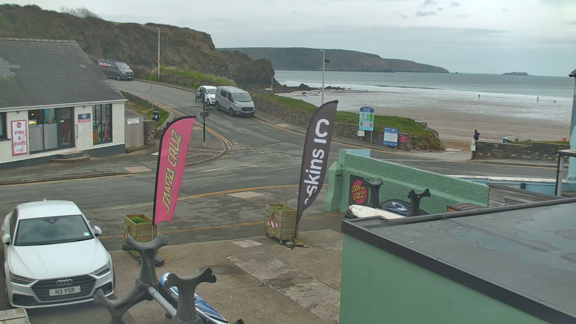A street scene overlooking a beach on an overcast day shows several vehicles parked and driving near a building with a fish and chips shop, with a sandy beach and calm ocean visible in the background.