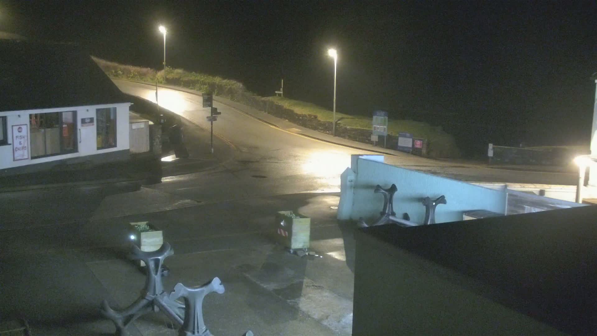 A wet, empty street at night curves past a small building, with two large anchors visible in the foreground.