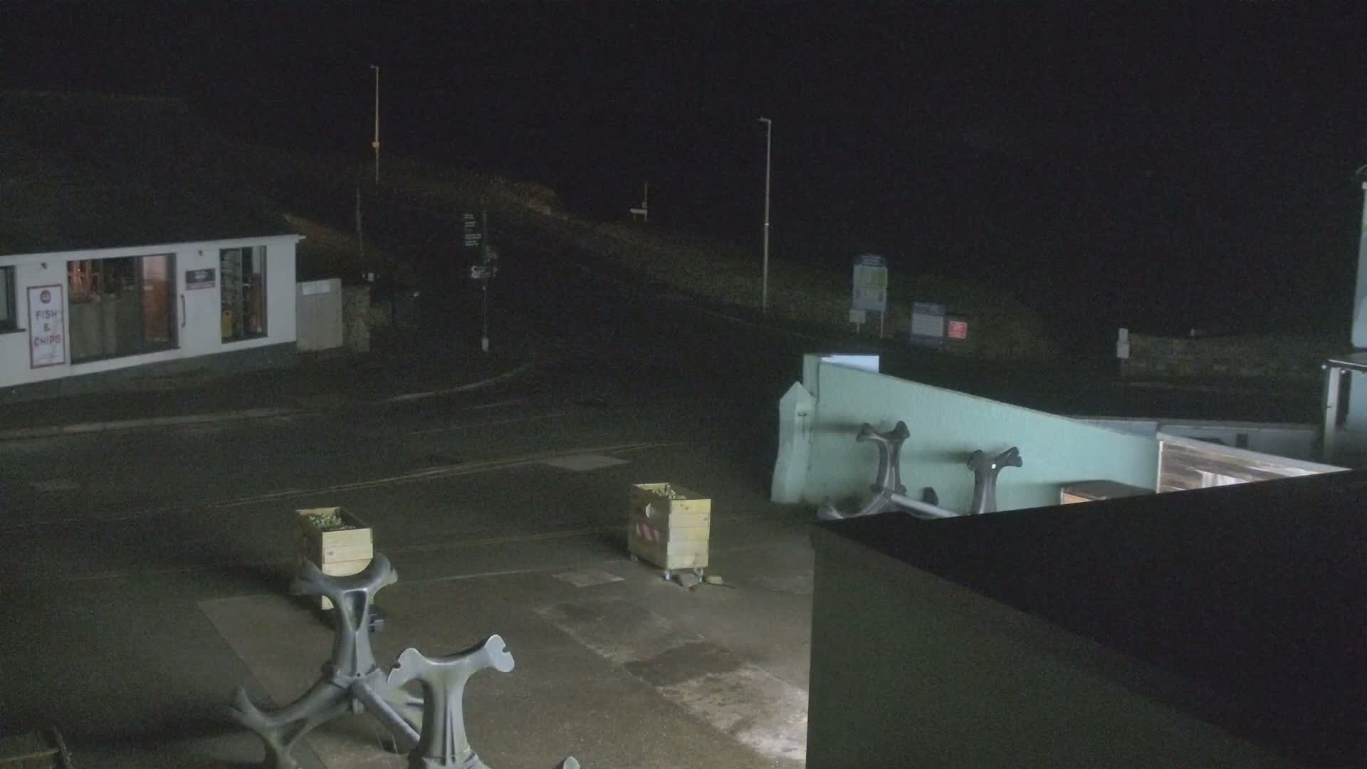 At night, a mostly empty street scene shows a small building, two wooden crates, and several dark metal fixtures in a parking lot.