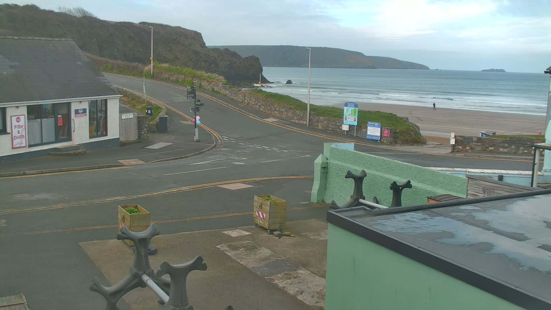 A coastal road curves around a building toward a sandy beach with small waves under an overcast sky.