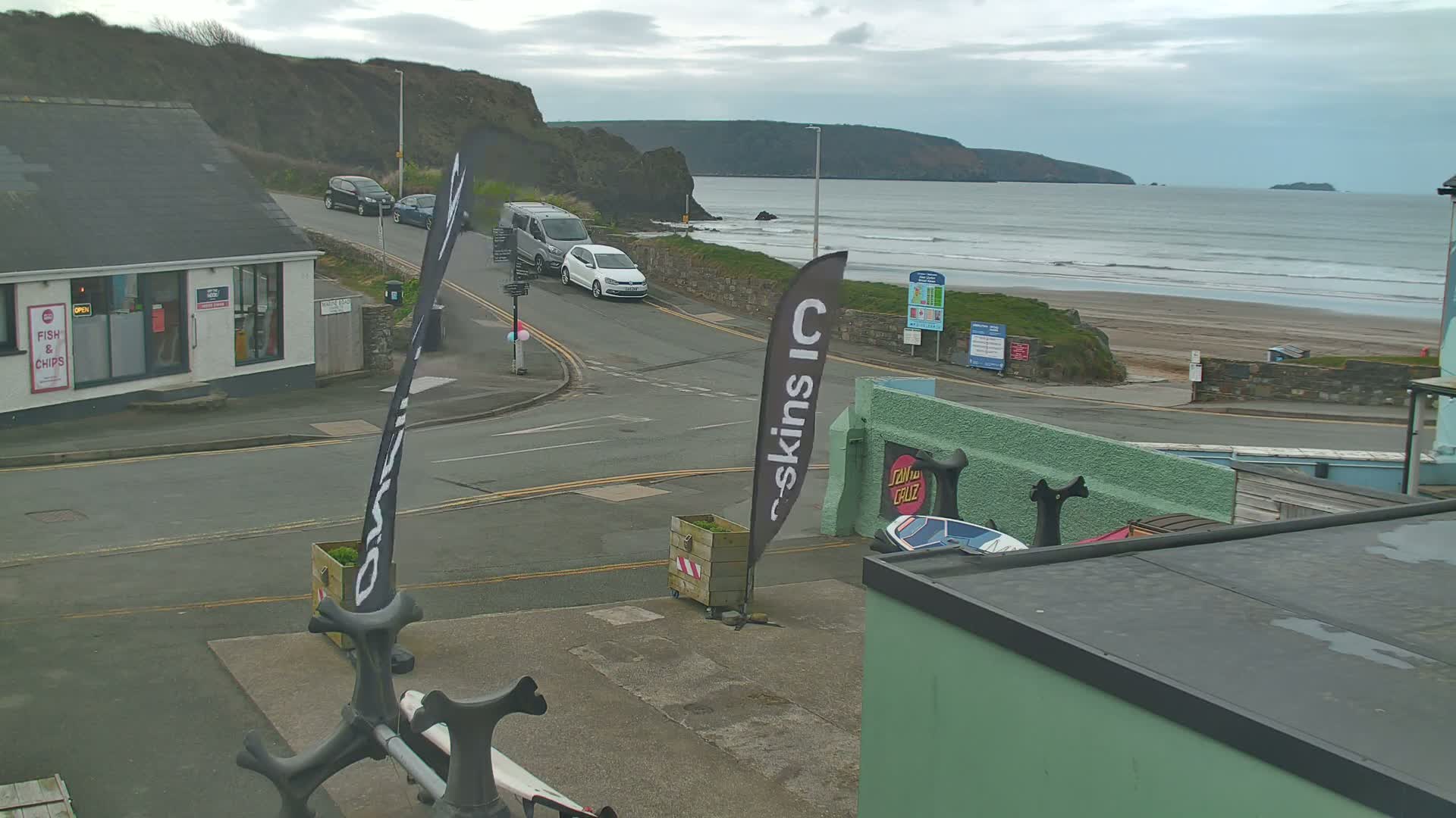 A coastal town scene on an overcast day shows a street intersection near a beach with gentle waves, a few cars, and buildings including a fish and chips shop.