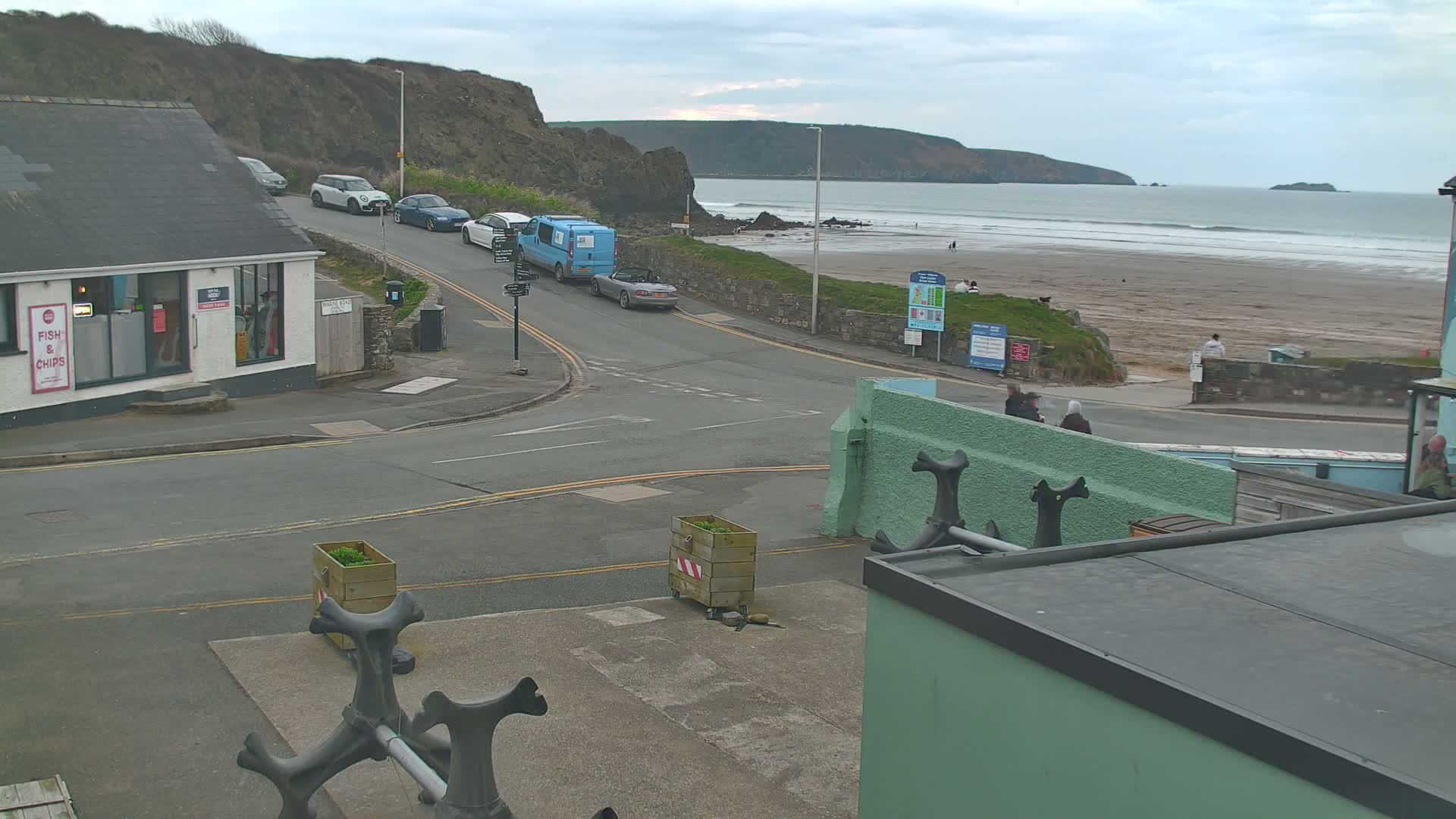 A coastal road intersects near a fish and chips shop, with several cars parked and driving, overlooking a sandy beach with gentle waves under an overcast sky.