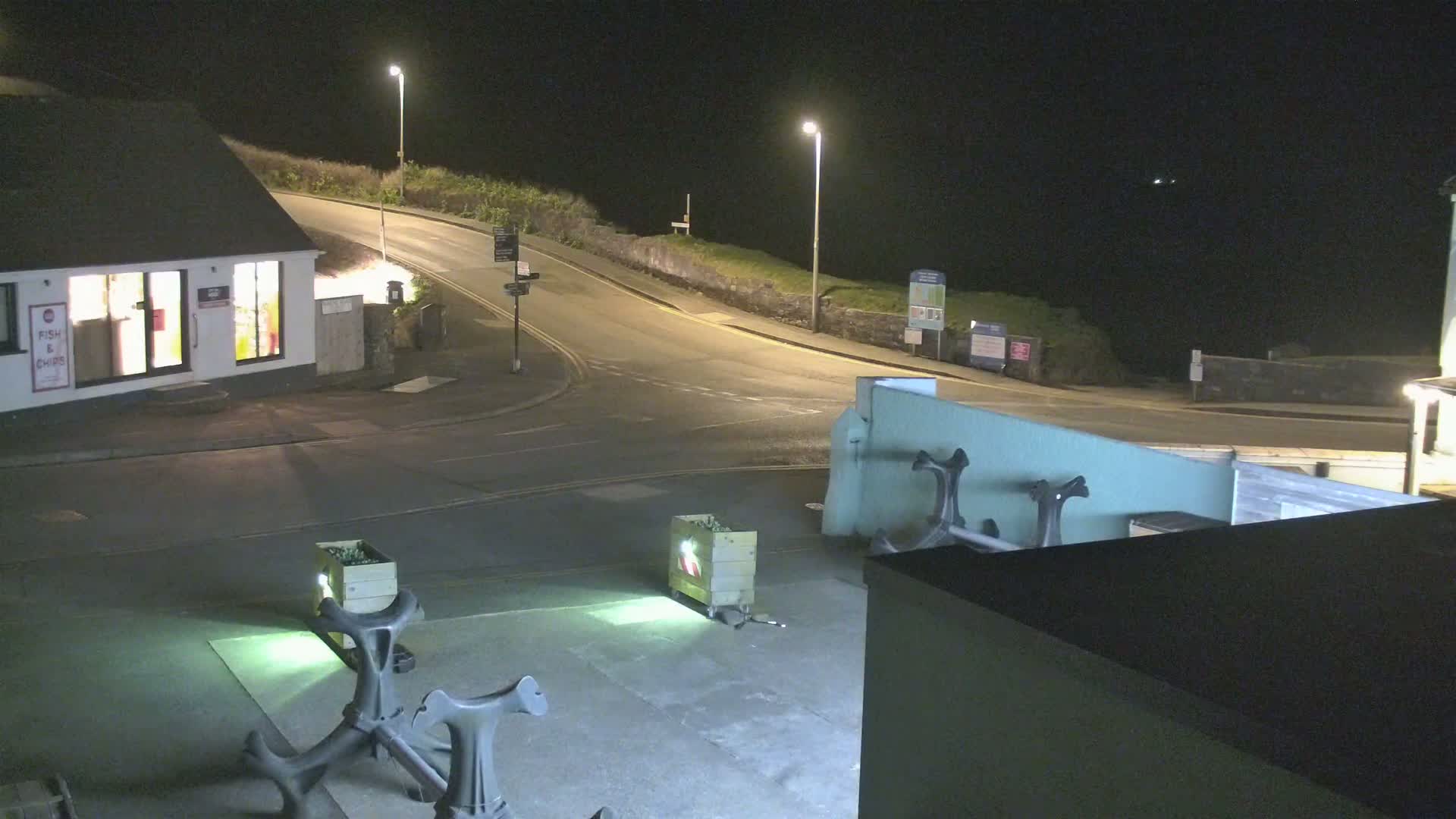 At night, a street with a fish and chips shop curves near a body of water, where two anchors and illuminated containers sit in a small plaza.
