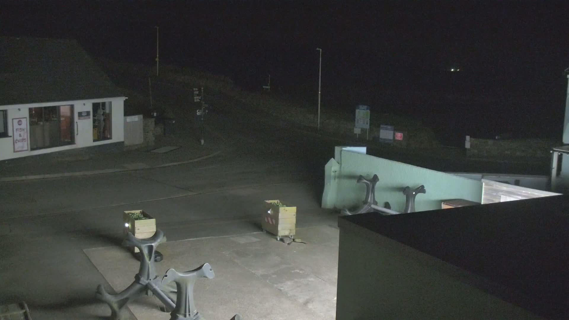 At night, a mostly empty paved area is visible, featuring a small building, several wooden crates, and some dark metal bollards; a faint light is seen in the far distance.