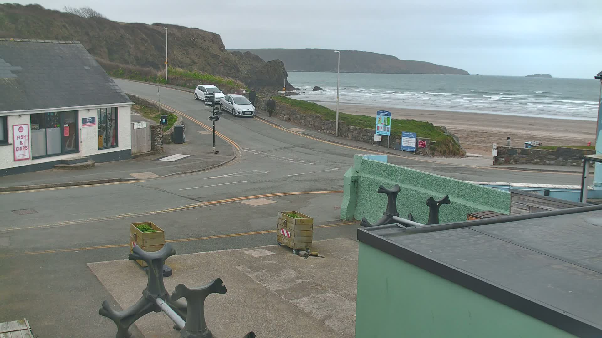 A street scene overlooking a beach with wavy ocean under an overcast sky, featuring parked cars, a small building, and decorative anchors.