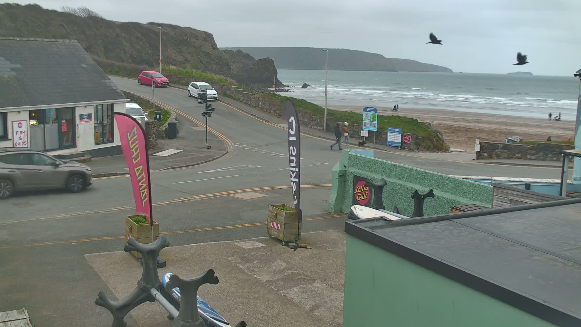 Overcast skies and a choppy ocean are visible beyond a road intersection near a beach, where a few people are walking and several cars are parked.