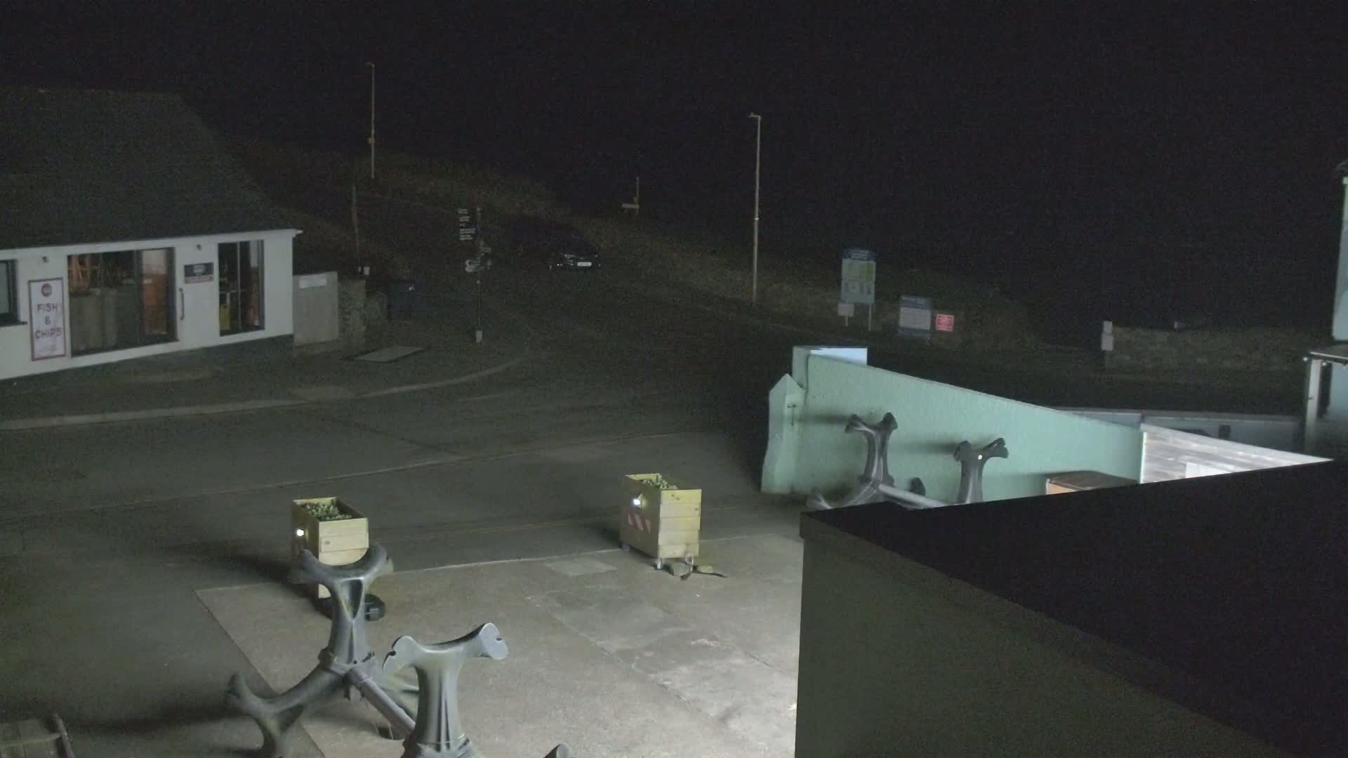 At night, a paved area with two wooden crates and several dark metal objects is illuminated, near a small building and a parked car under a dark sky.