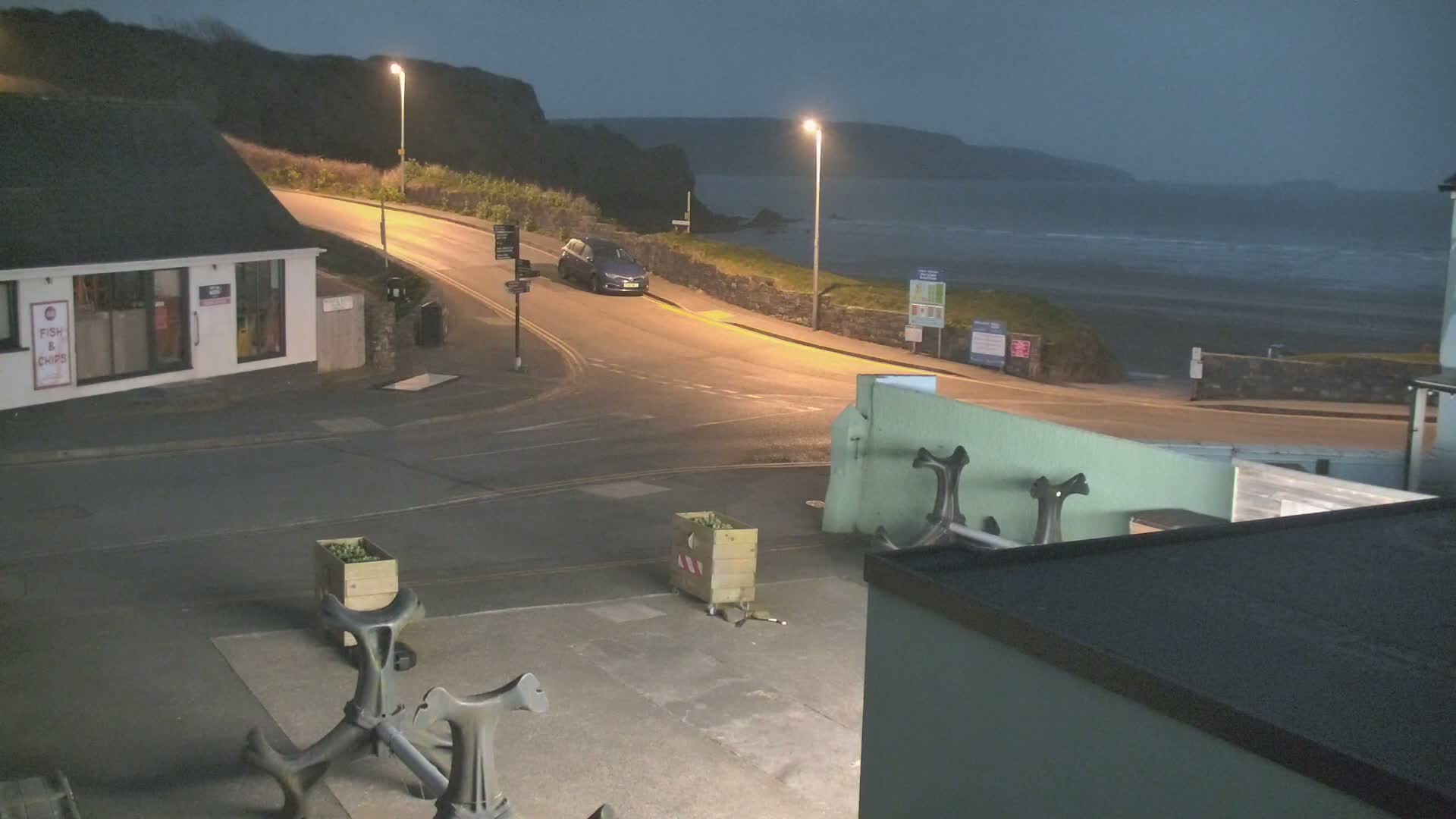 A dark, seaside town at night with a car parked on a curving road overlooking a wavy ocean under a dim sky.