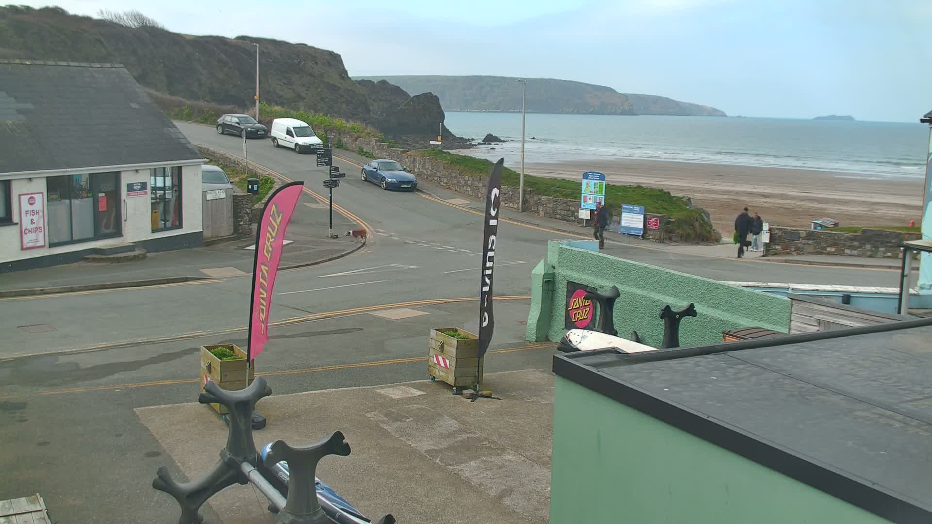 A street scene overlooking a beach on an overcast day shows several cars parked and driving near a building; some pedestrians are walking along the beach and road.