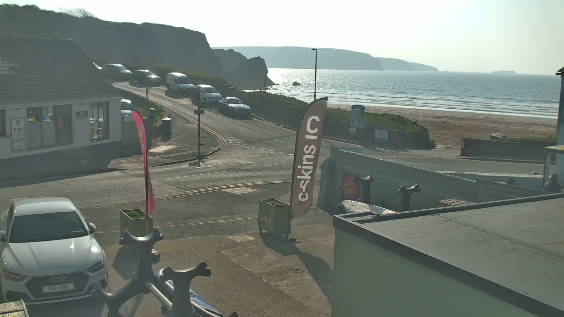 A sunny seaside town scene shows a road curving toward a sandy beach with a calm ocean under a hazy sky, several cars parked along the roadside, and a building with a flat roof.