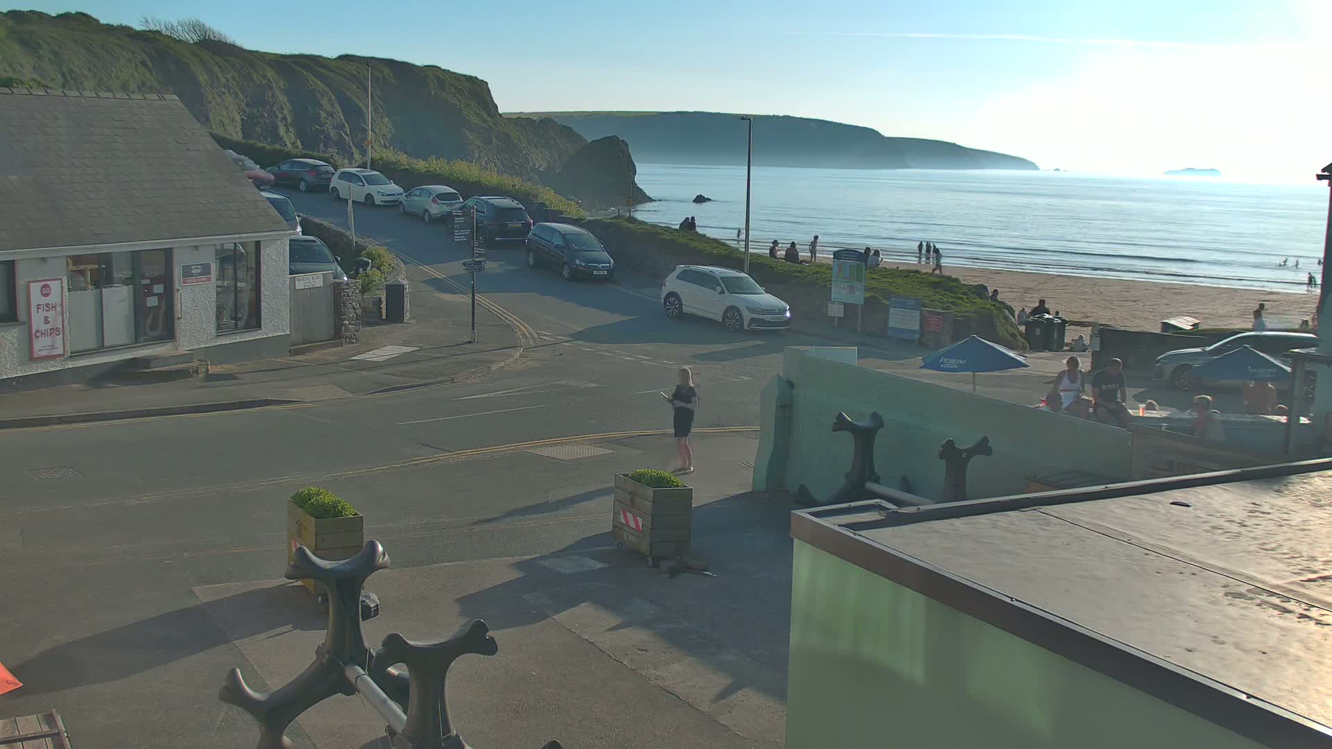 A sunny seaside town scene shows cars parked along a road leading to a beach with people relaxing on the sand and in the water, under a mostly clear sky.