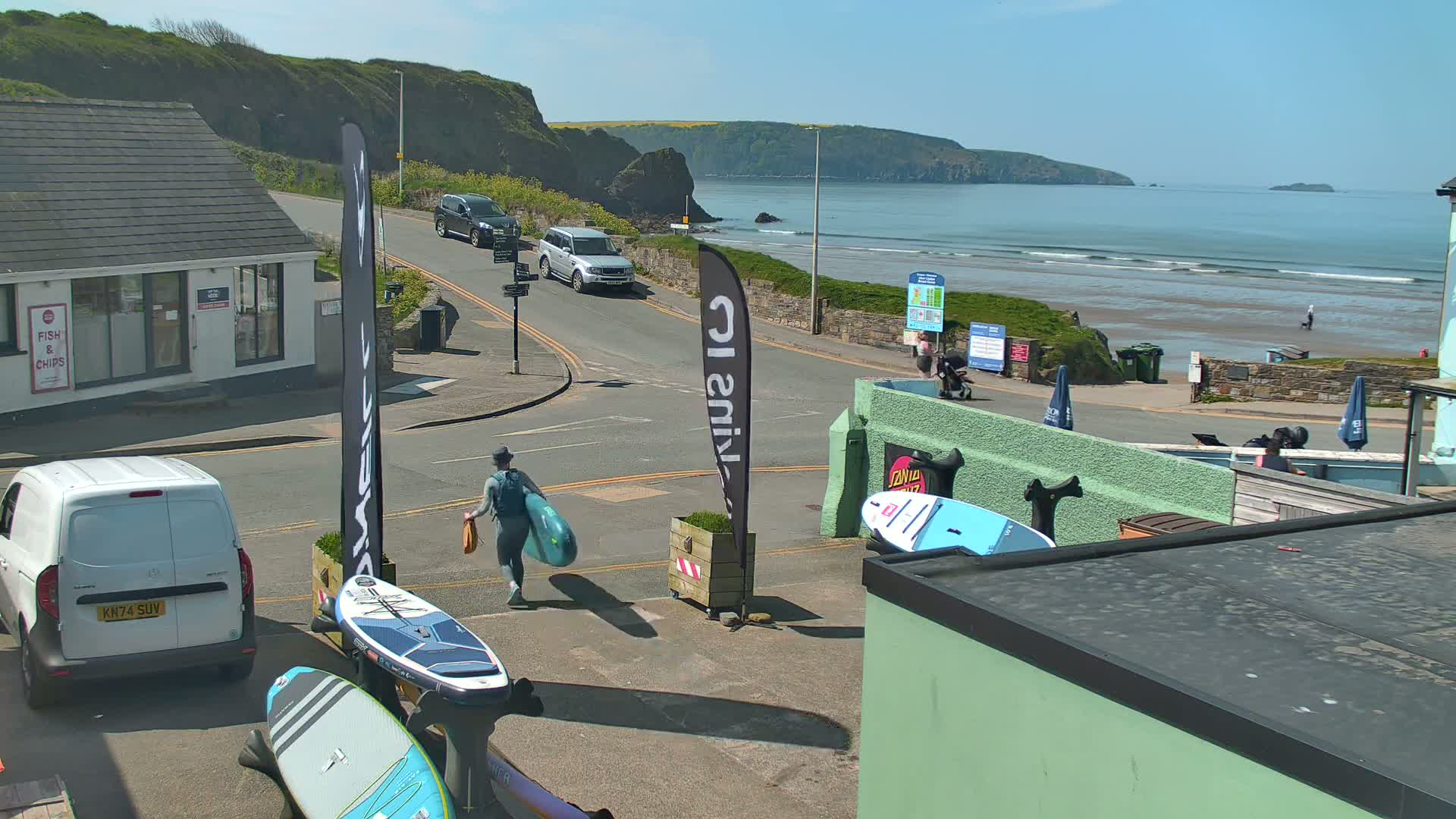 A person carries a paddleboard down a street toward a beach with gentle waves on a sunny day.