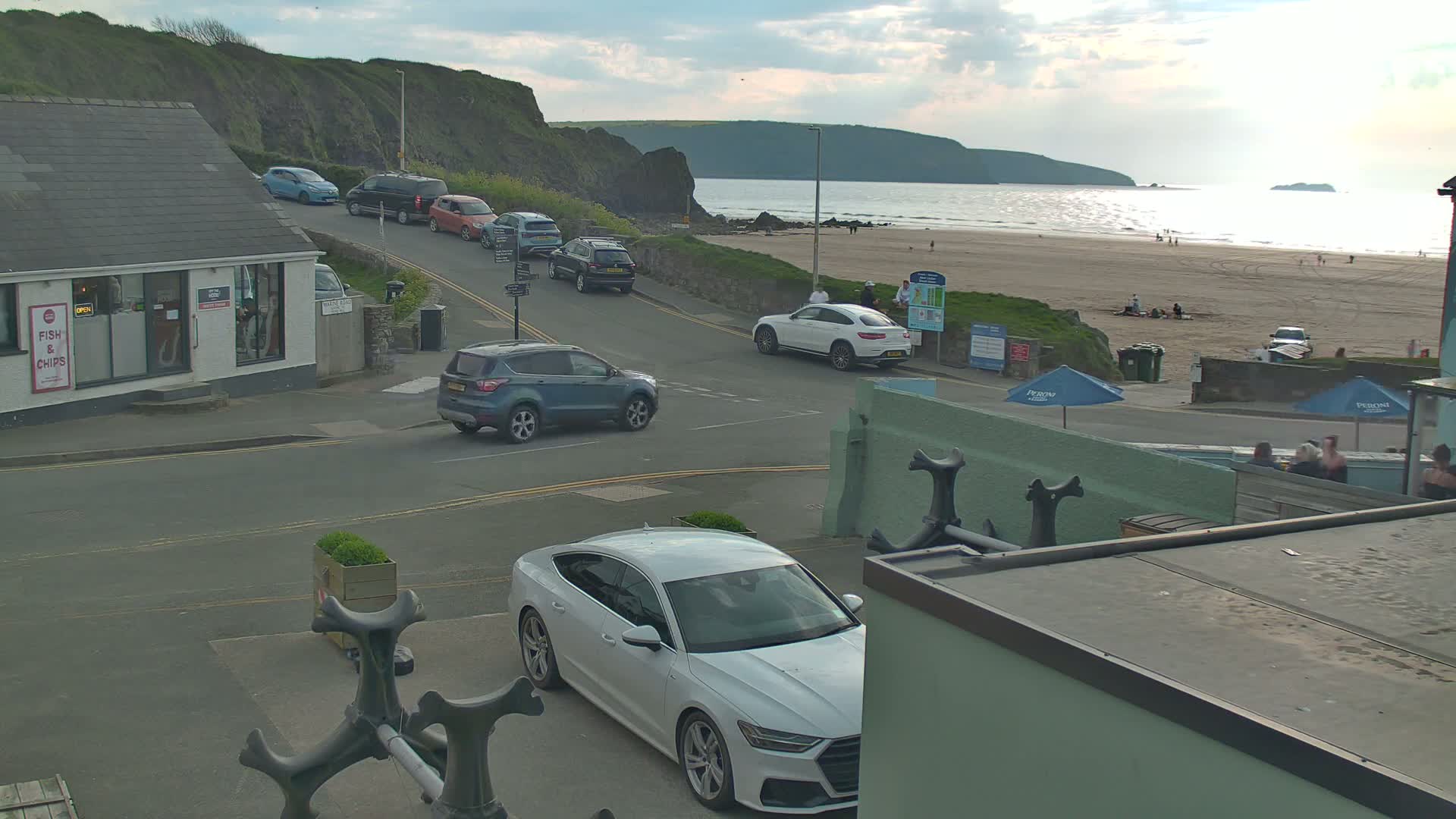 A coastal town scene shows several cars parked and driving on a street near a sandy beach under a partly cloudy sky.