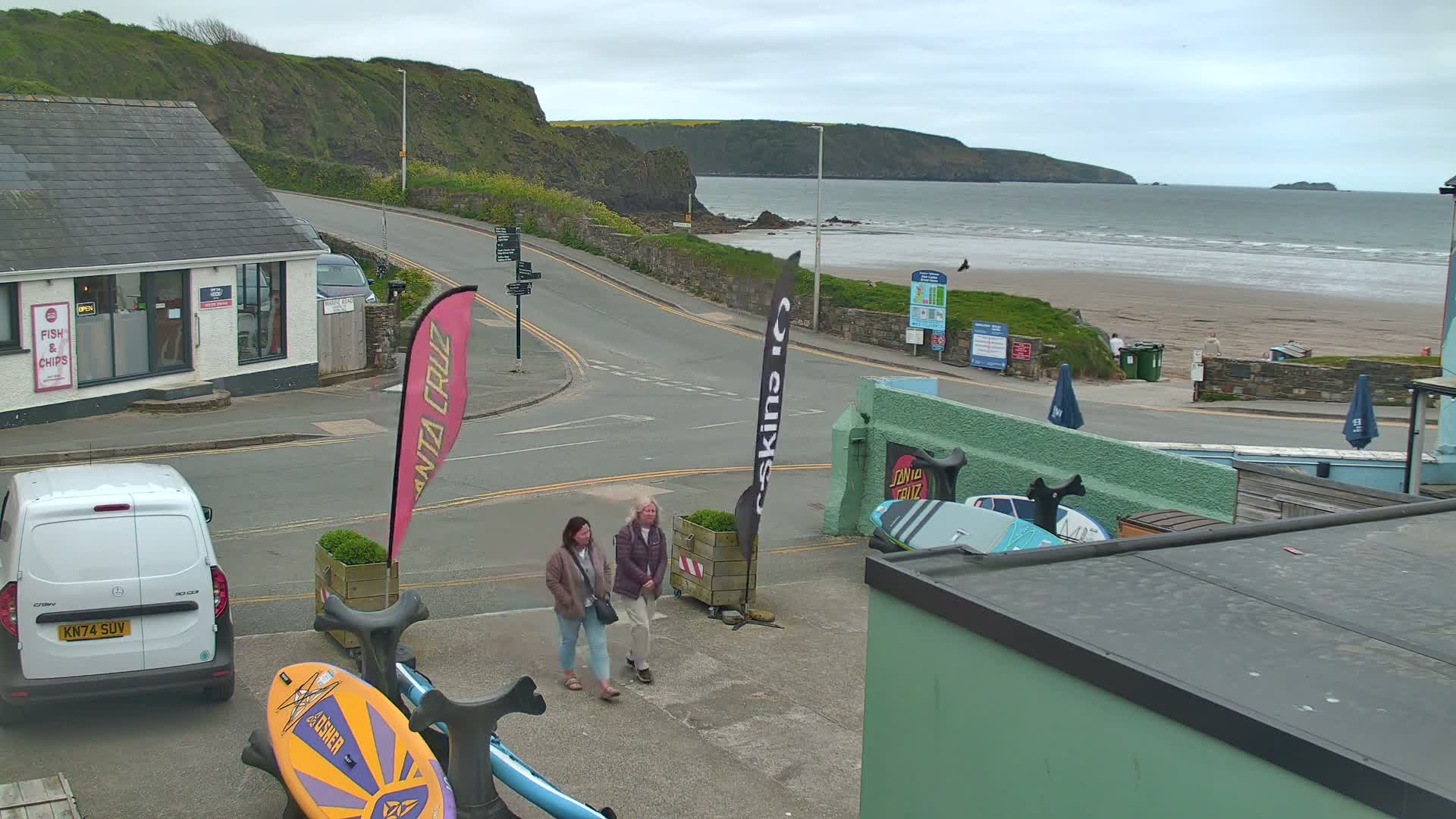 Two people walk past a seaside parking lot with surfboards, a van, and a building near a sandy beach under an overcast sky.
