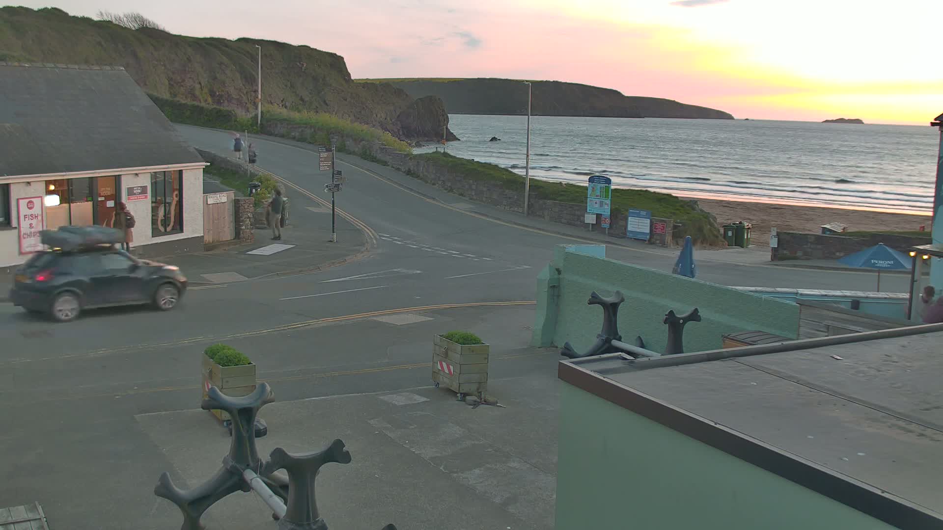A seaside town at sunset, with a car parked near a fish and chips shop, overlooks a beach with gentle waves.