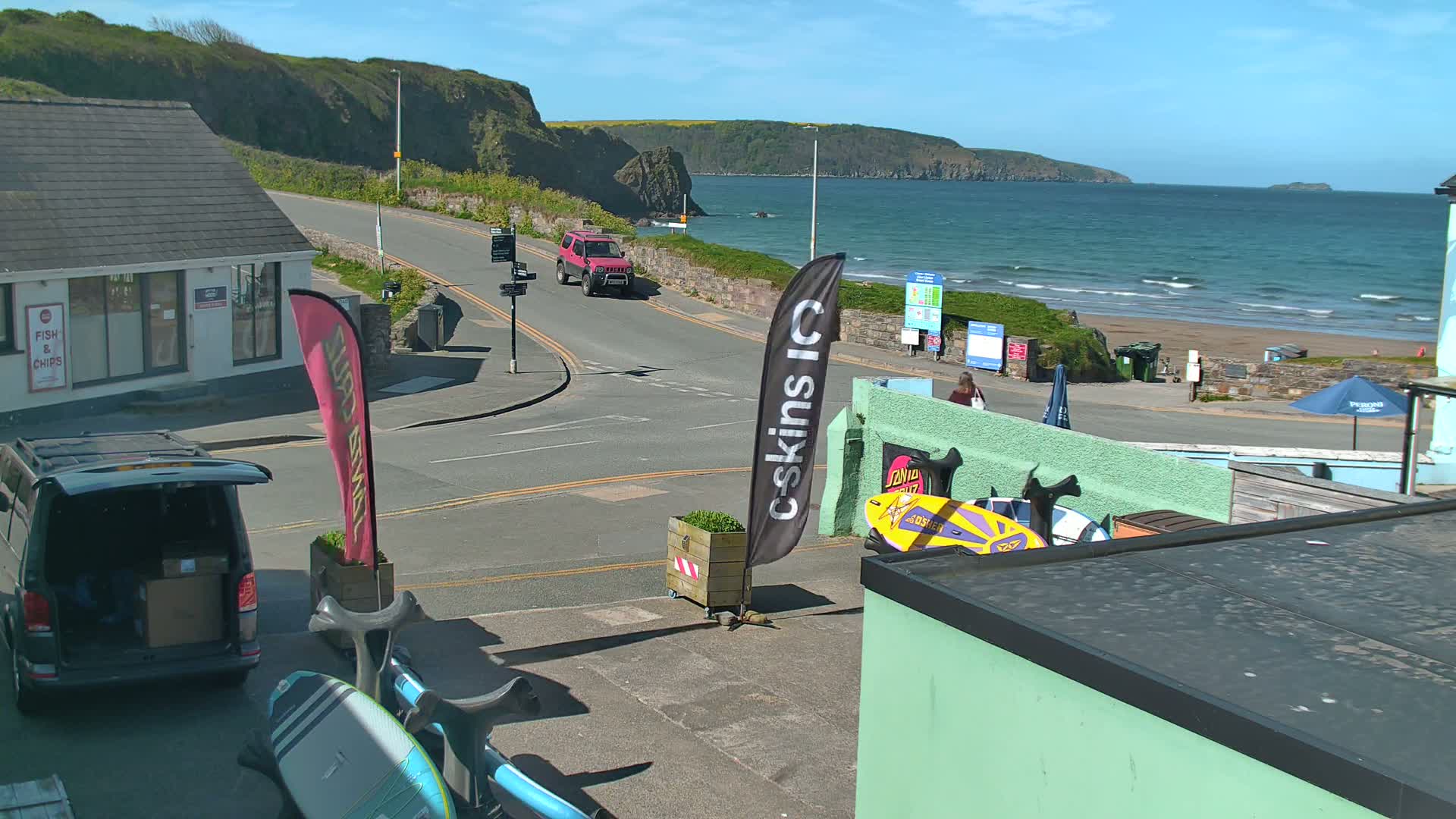 A sunny seaside town scene shows a road leading to a sandy beach with small waves, a red SUV driving down the road, and surfboards leaning against a building.