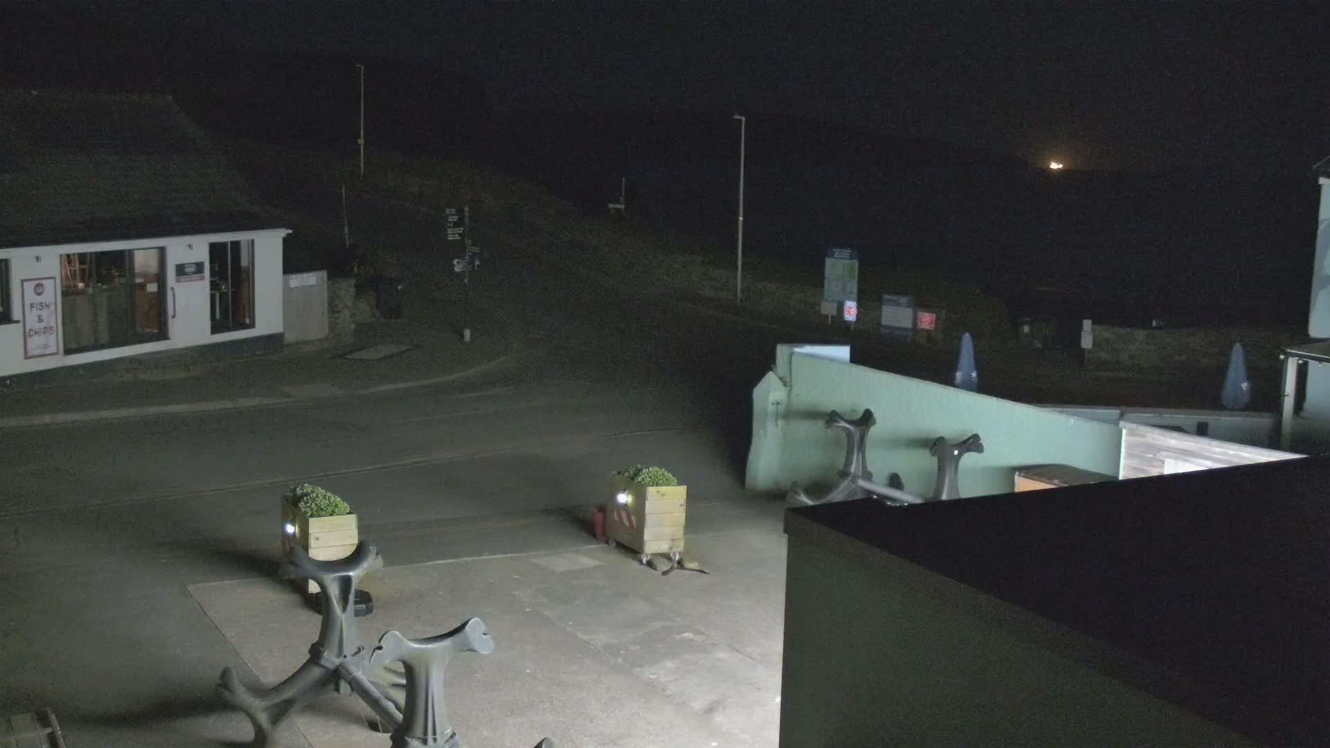 At night, a small town square with a fish and chips shop is illuminated, showing several decorative anchors and planters, with a dark, possibly cloudy sky visible in the distance.