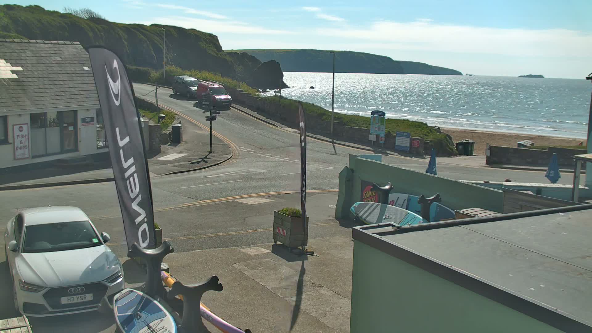 A sunny coastal scene shows a road leading to a beach with calm, sparkling water under a mostly clear sky, with parked cars and a few buildings visible.