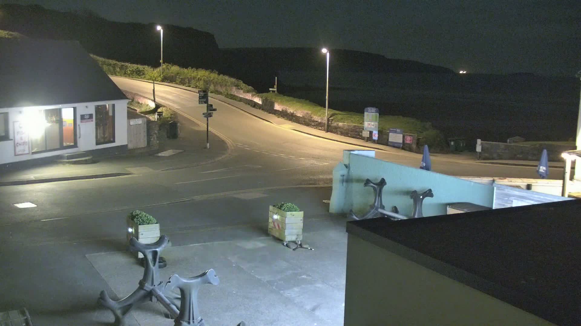 At night, a coastal road curves around a building and toward a dark, cliff-lined ocean.