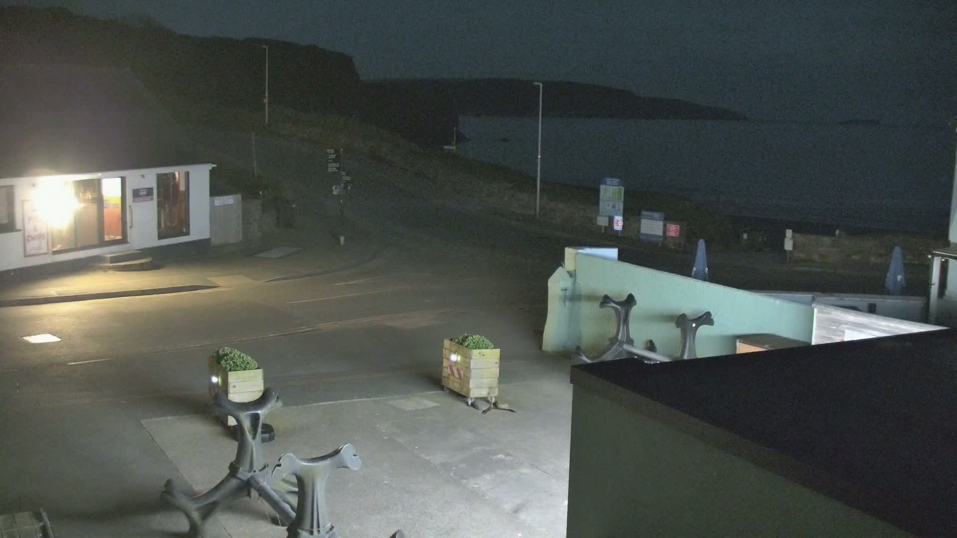 At night, a dark-colored body of water is visible beyond a small building and an empty paved area with several anchors and planters.