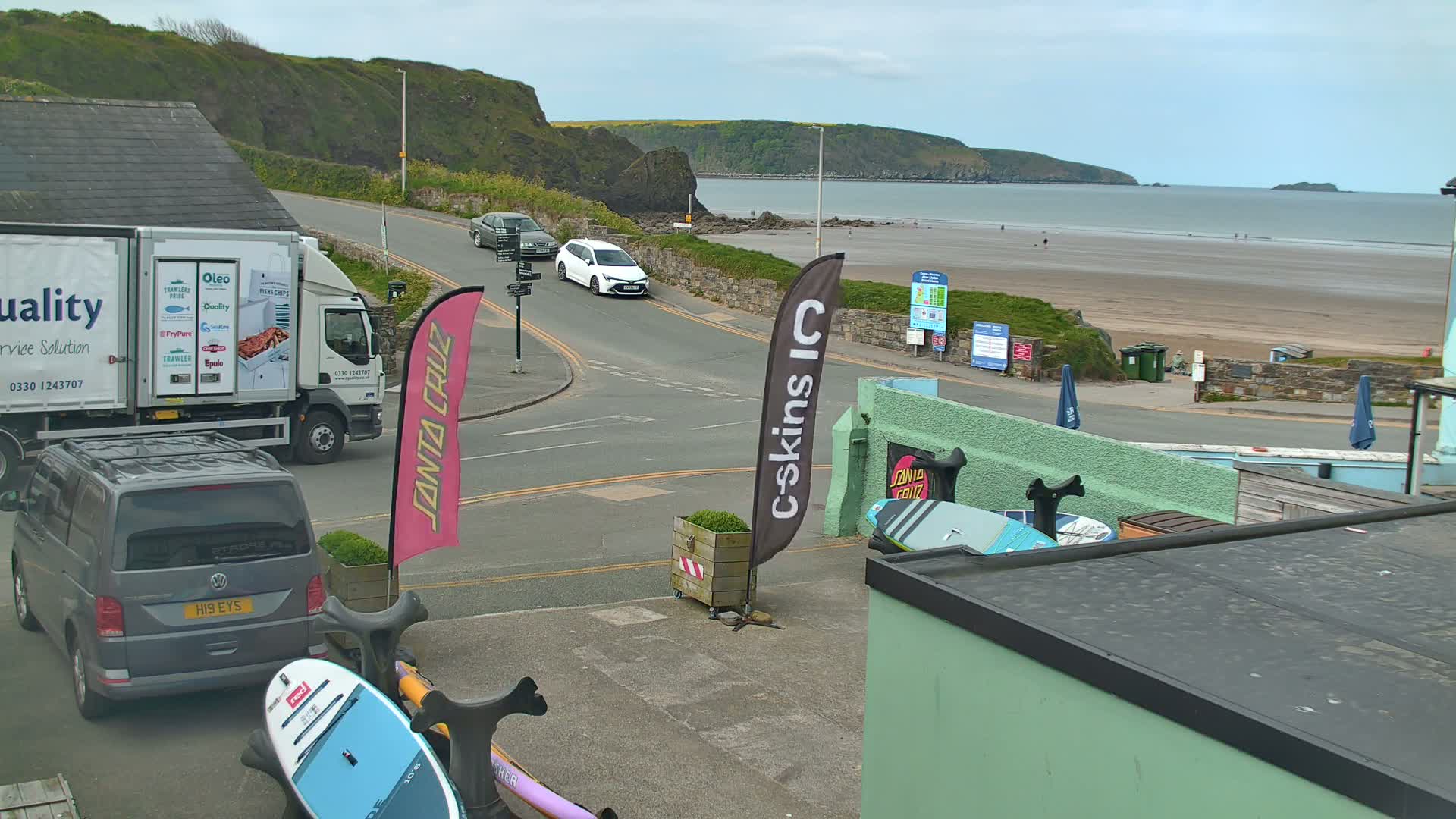 A street scene overlooking a sandy beach on a cloudy day shows a delivery truck, cars, and paddleboards near a building.
