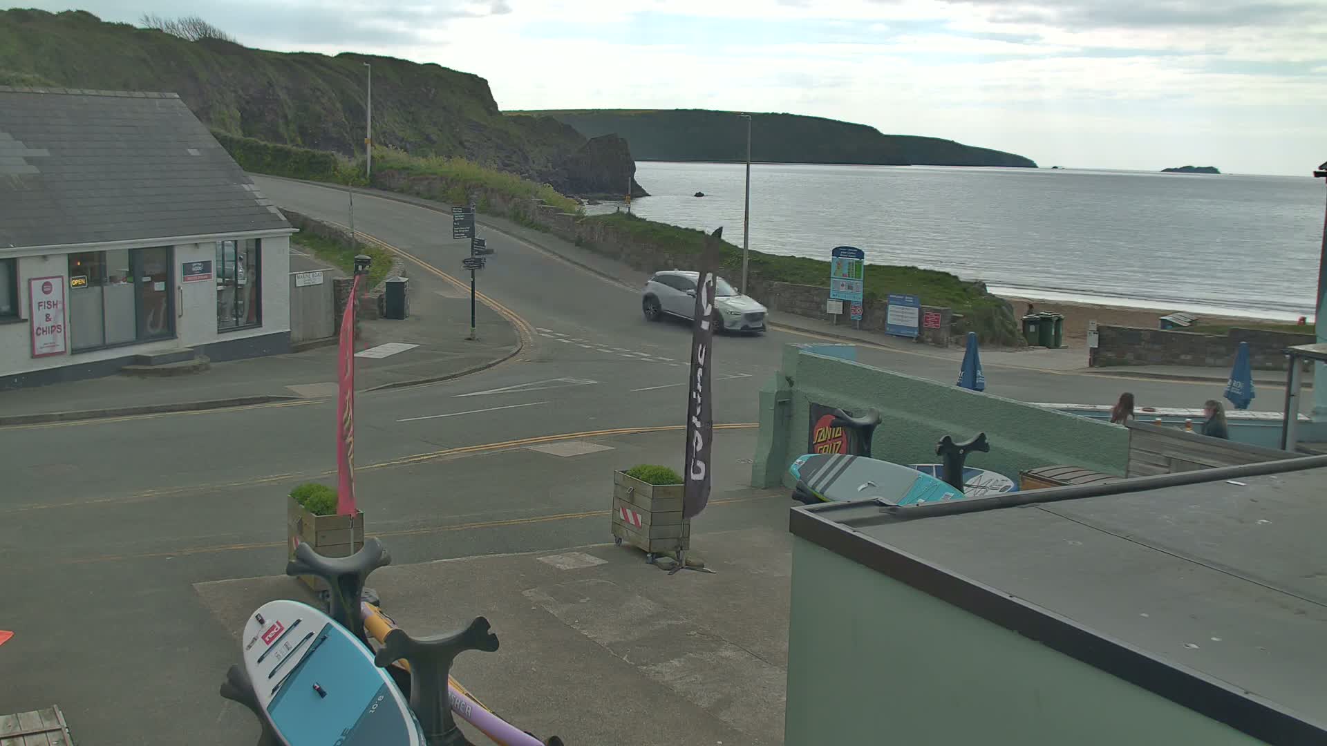 A seaside town scene on an overcast day shows a road curving toward a beach with a calm sea and distant hills, paddleboards are visible near a building.