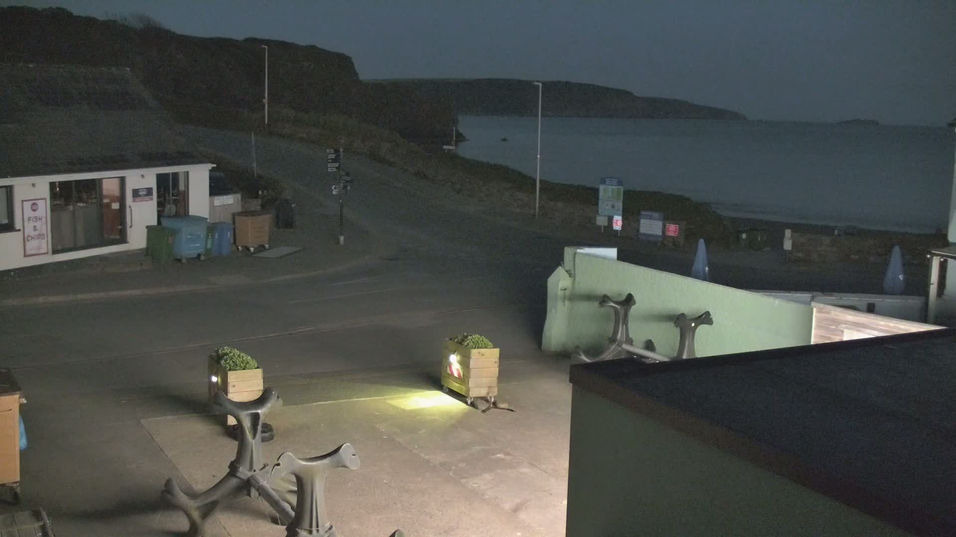 A nighttime coastal scene shows a dark, calm sea under a dusky sky, with a small building, several planters, and anchors visible in the foreground.