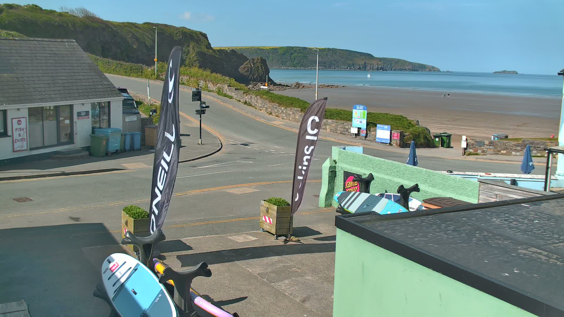 A sunny seaside view shows a road leading to a sandy beach with a few people visible, alongside buildings, and several paddleboards displayed near a street corner.