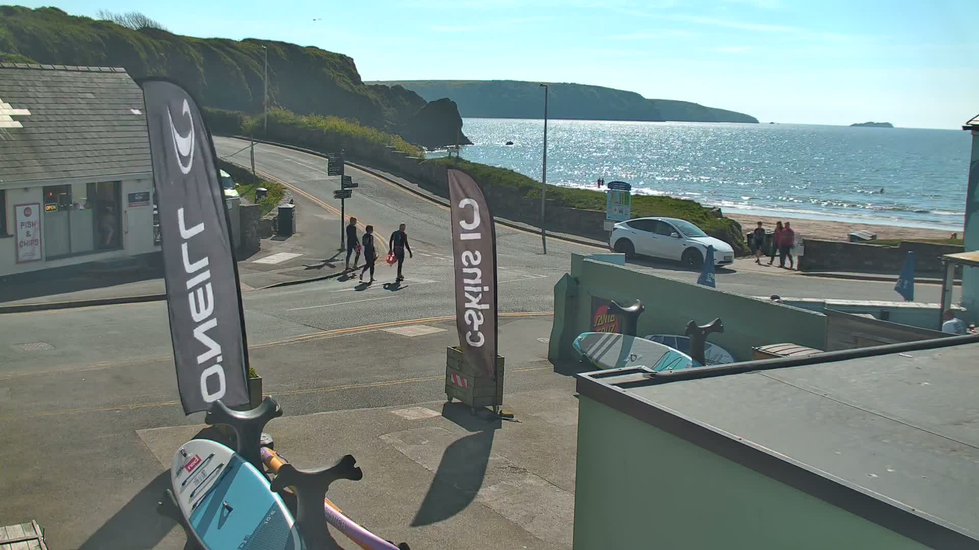 A sunny seaside scene shows a street with people walking, a white car parked, and surfboards leaning against a building near a beach with calm waves.
