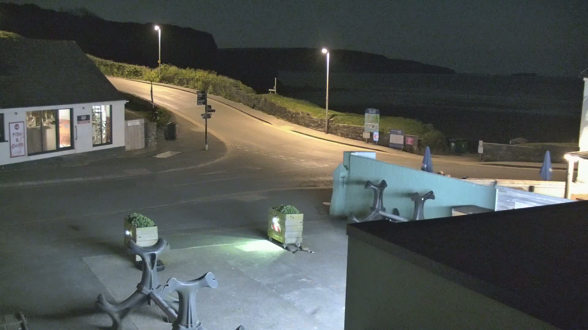 At night, a coastal road curves around a building toward a dark body of water under illuminated streetlights.