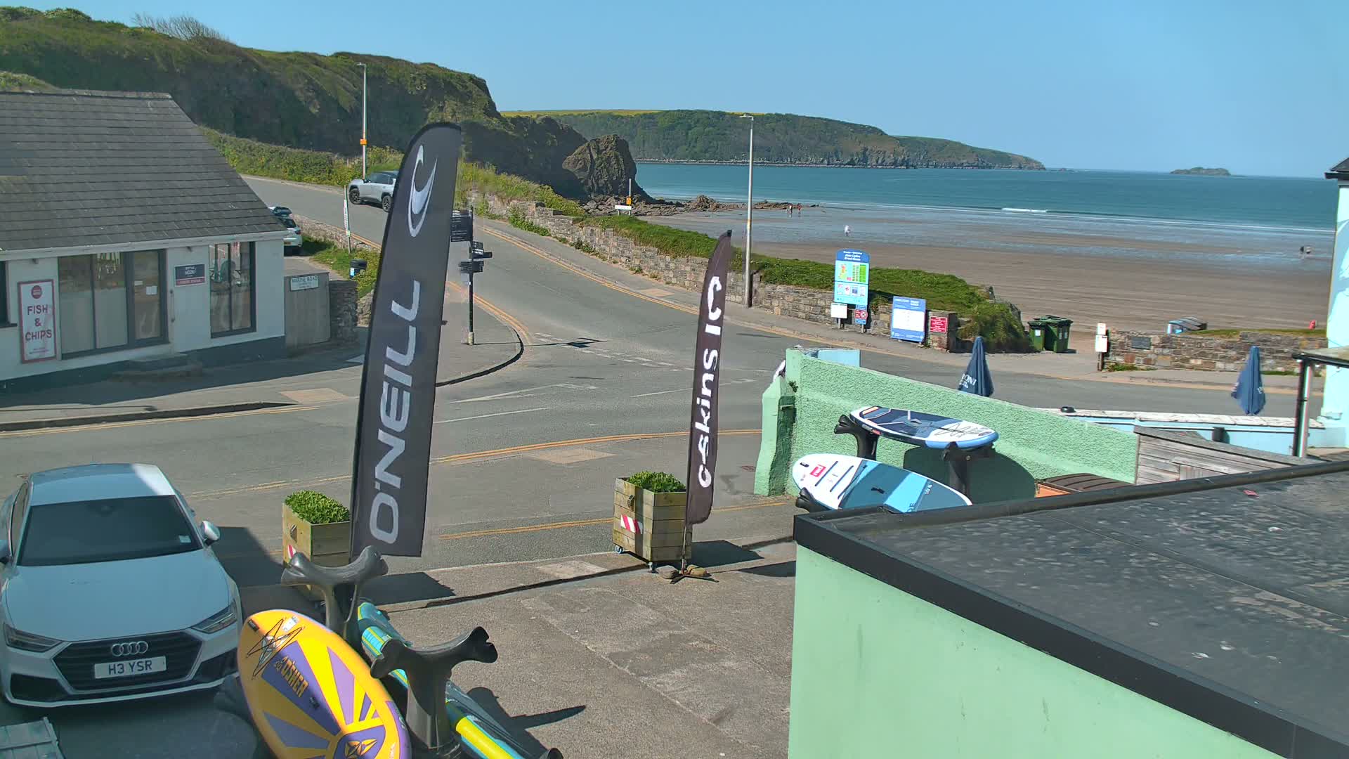 A sunny seaside town scene shows a street with parked cars, surfboards leaning against a building, and a sandy beach with a few people visible in the distance.