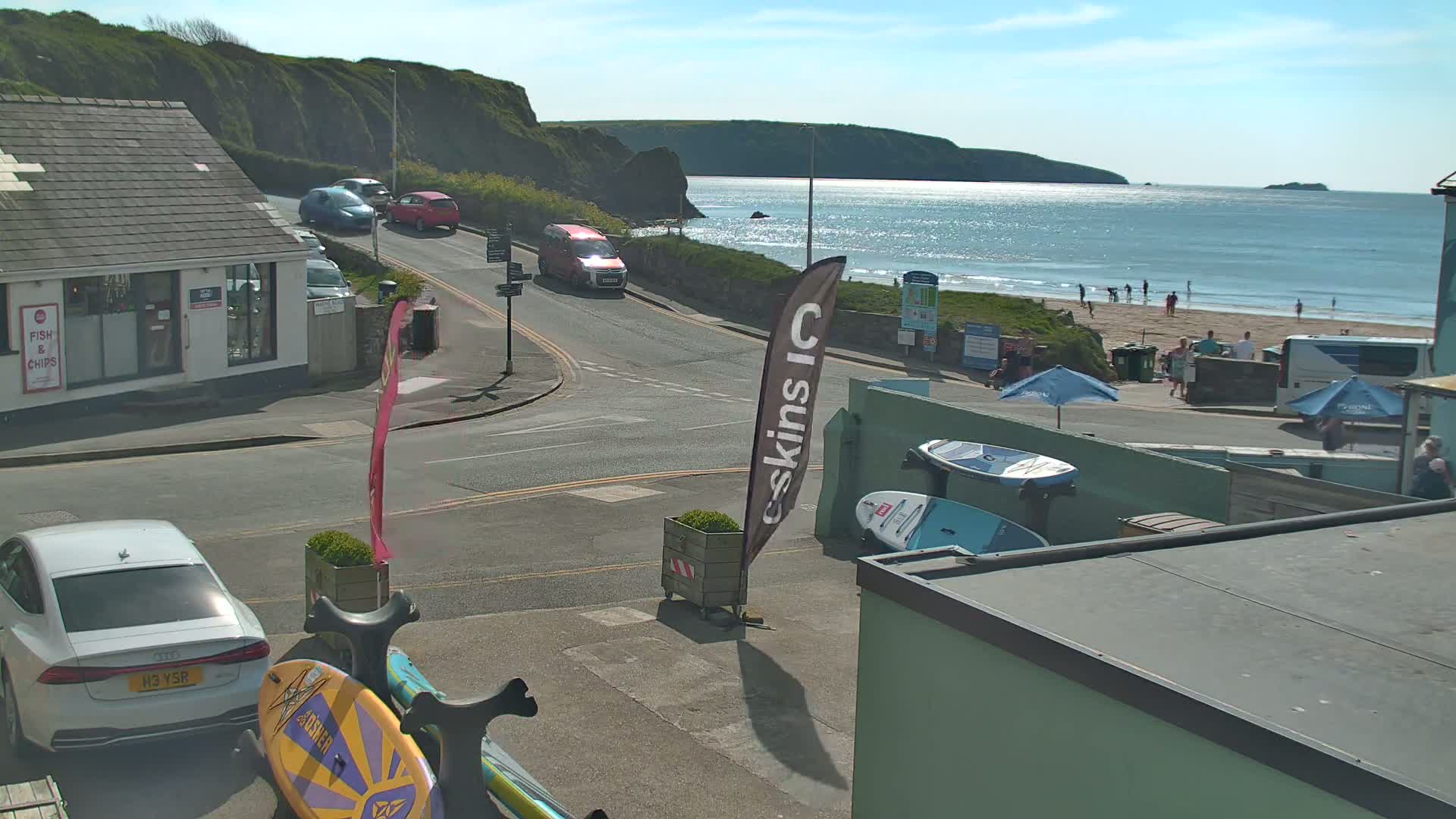 A sunny seaside town scene shows a street with parked cars curving toward a beach with people, a calm ocean, and cliffs in the background.
