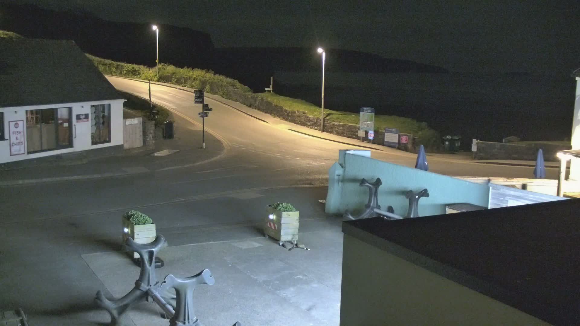 At night, a coastal road curves around a building and near the ocean, with several decorative anchors and planters on the ground.