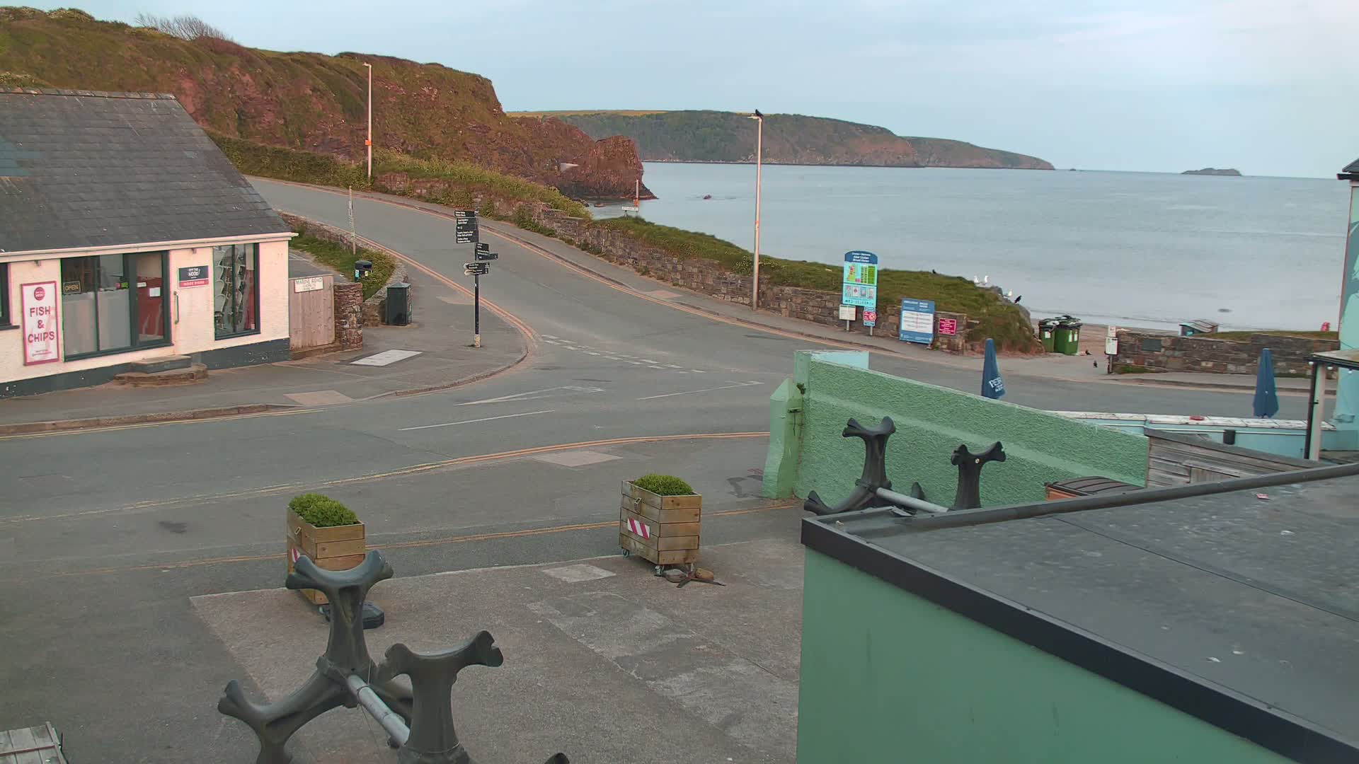 A coastal road curves around a small building and toward a calm sea under an overcast sky.