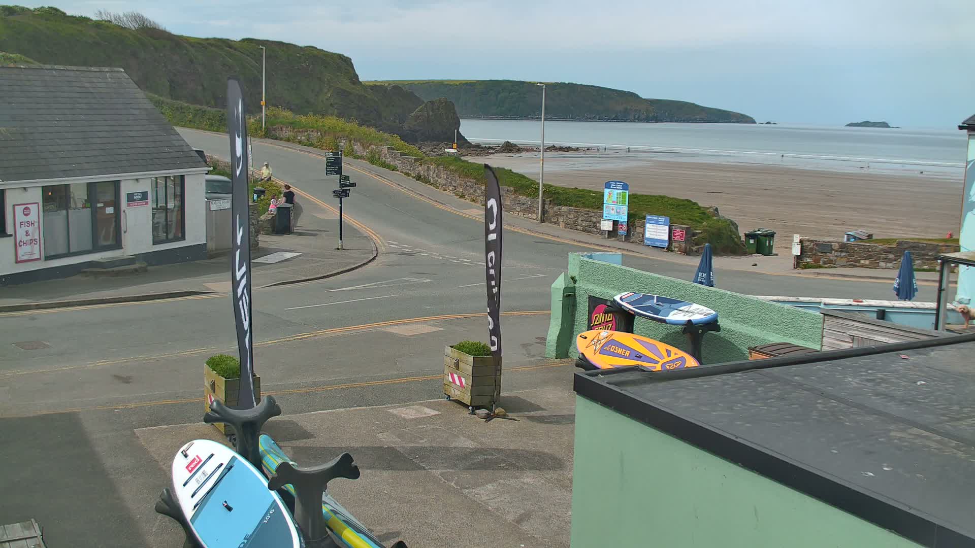 A sunny day overlooking a sandy beach with calm water, a road leading to it, and paddleboards stored outside a building.