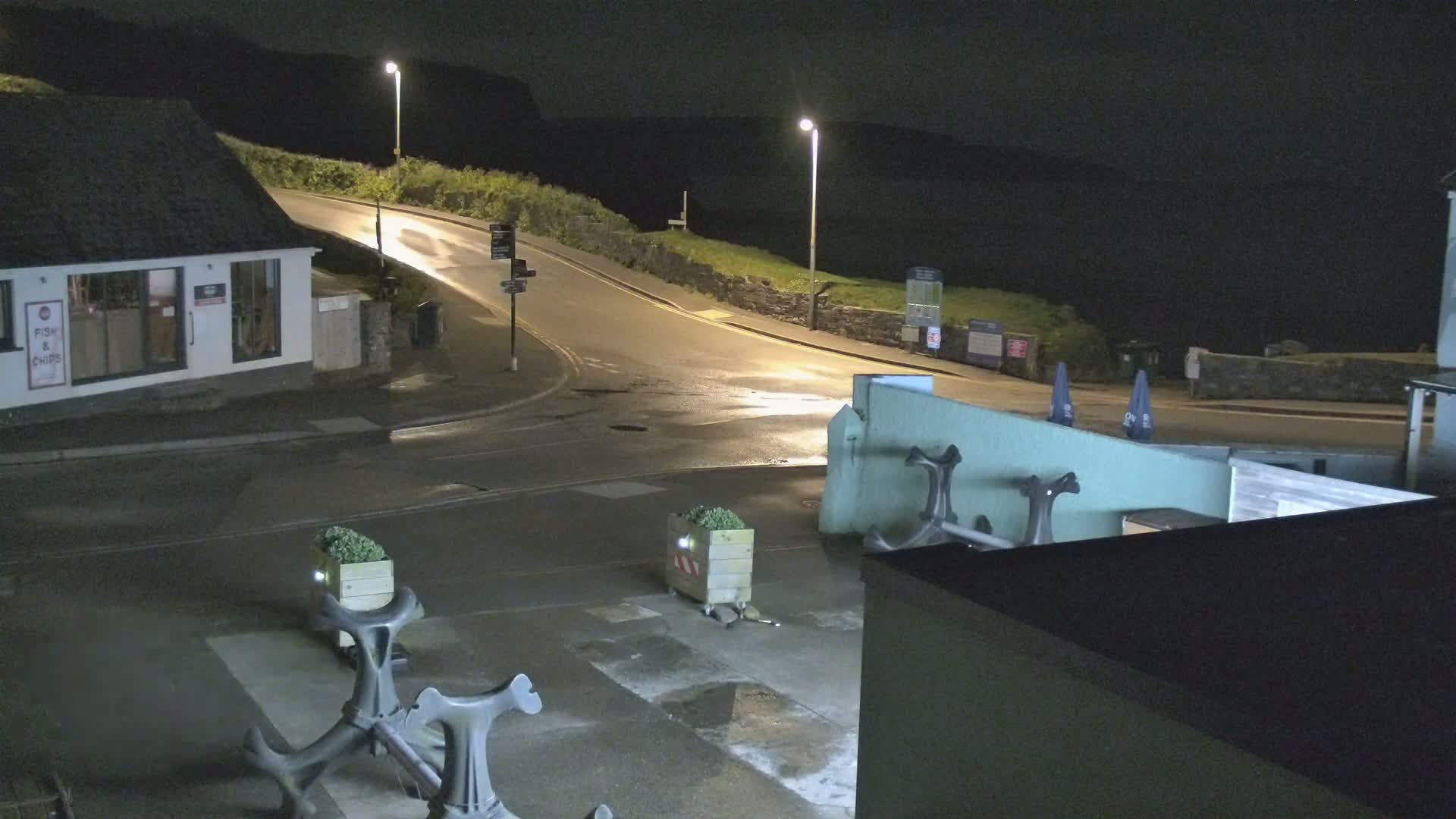 At night, a coastal road curves around a building with decorative anchors and planters, under illuminated streetlights.