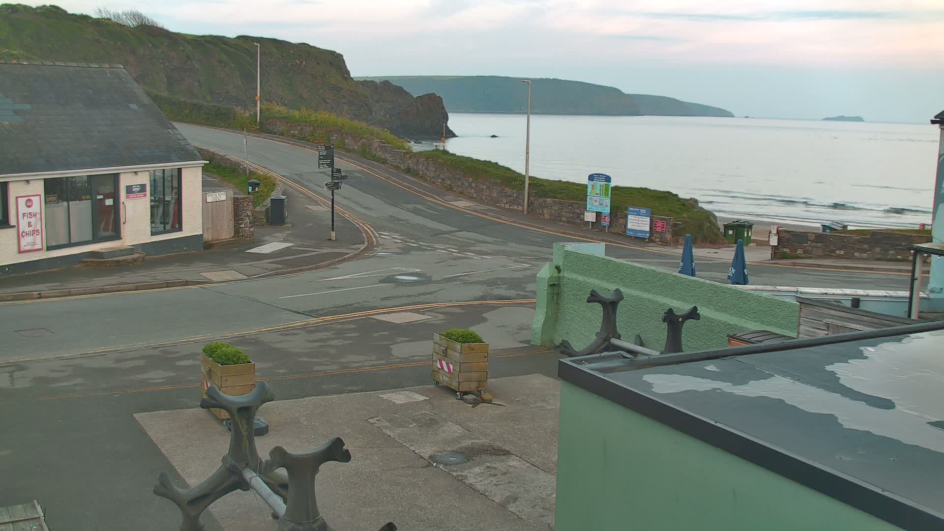 A coastal road curves around a cliffside, leading to a beach with gentle waves under an overcast sky.