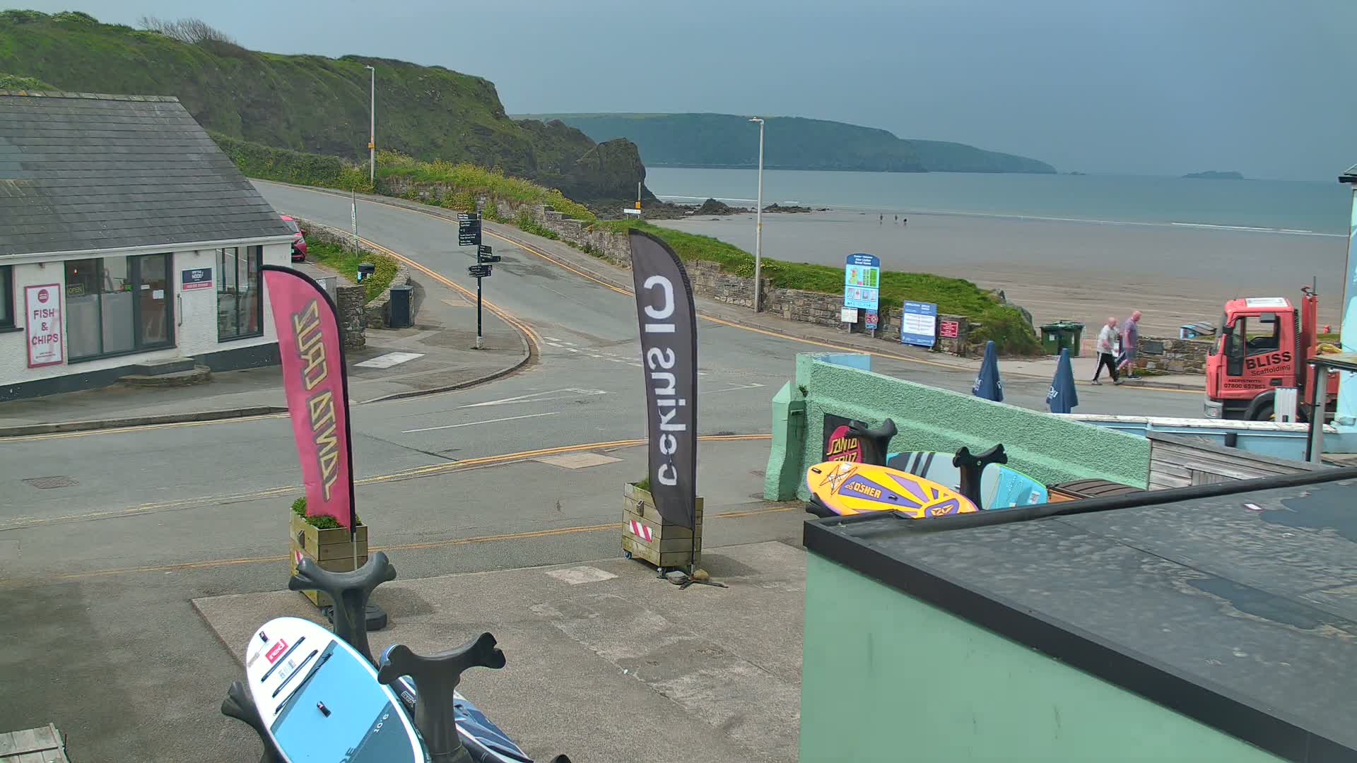 Overlooking a coastal road and beach on an overcast day, stand several surfboards near a building.