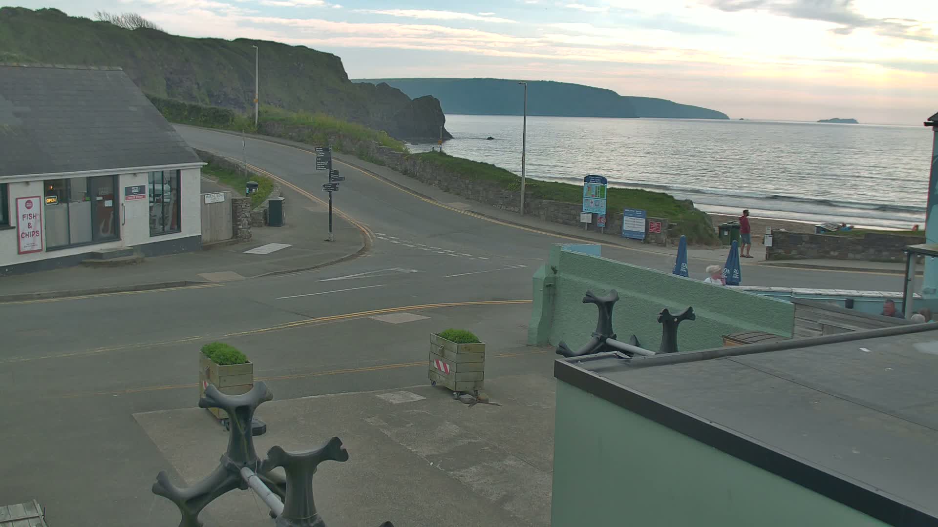 A coastal road curves around a seaside town, with a calm ocean and sandy beach visible under an overcast sky.