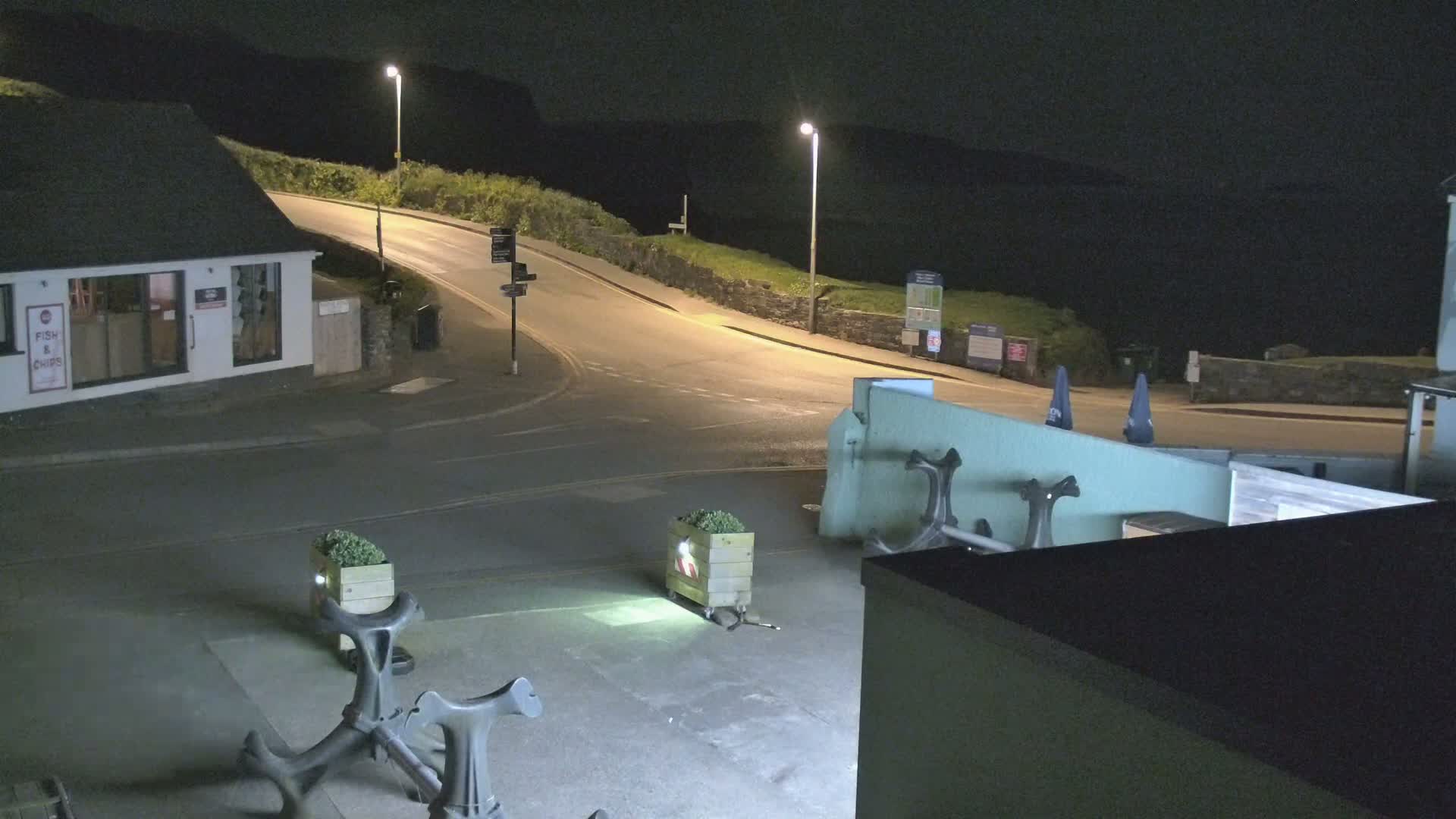 At night, a street corner scene shows a small building, a curving road leading uphill, and several planters and anchors in a paved area.