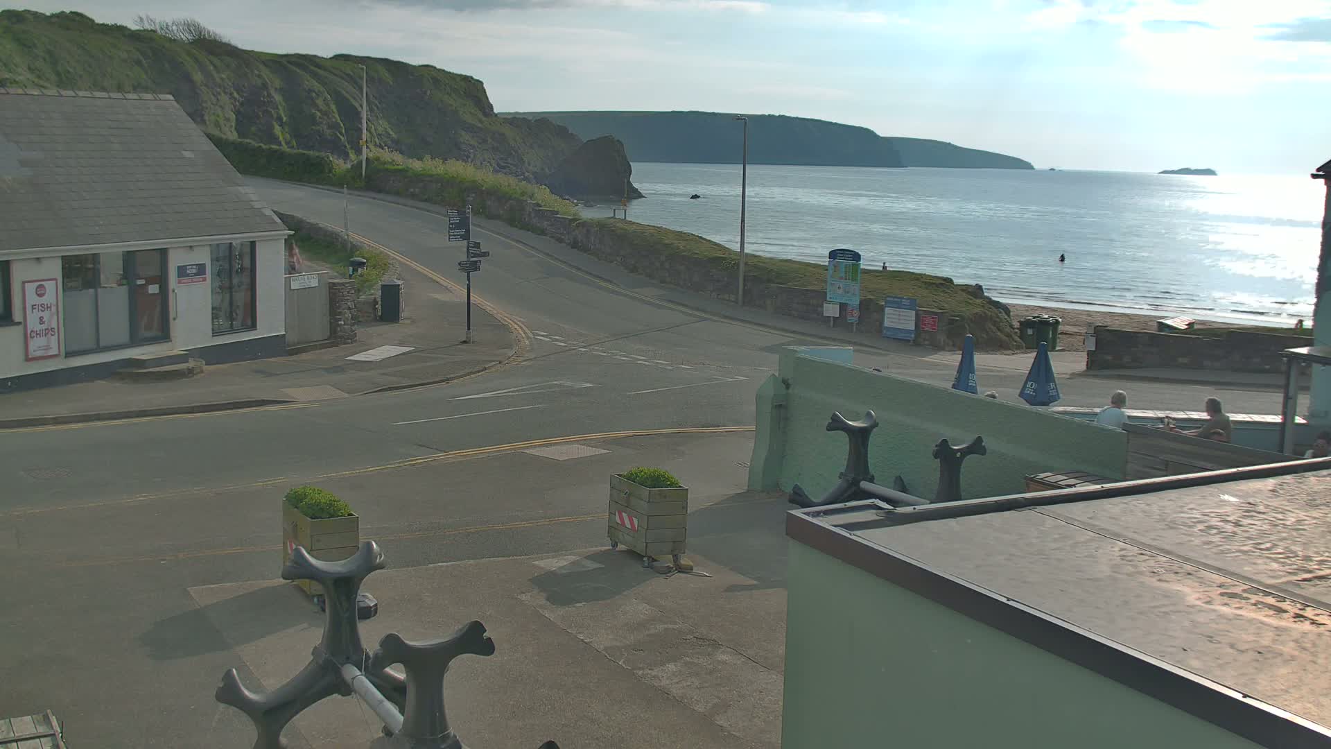 A sunny seaside town scene shows a road curving toward a beach with calm ocean water, a rocky cliff in the background, and several people relaxing near the shore.