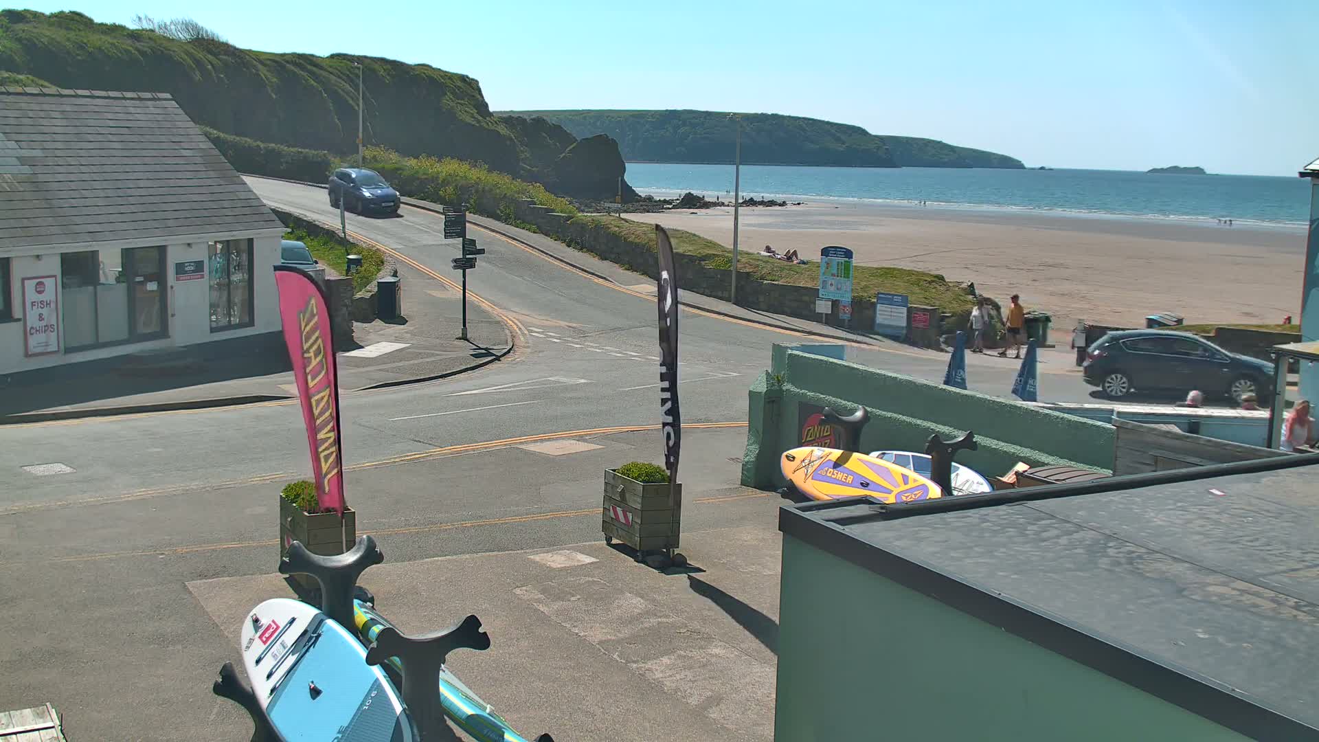 A sunny coastal scene shows a road leading to a sandy beach with people and parked cars, paddleboards are visible near a building.