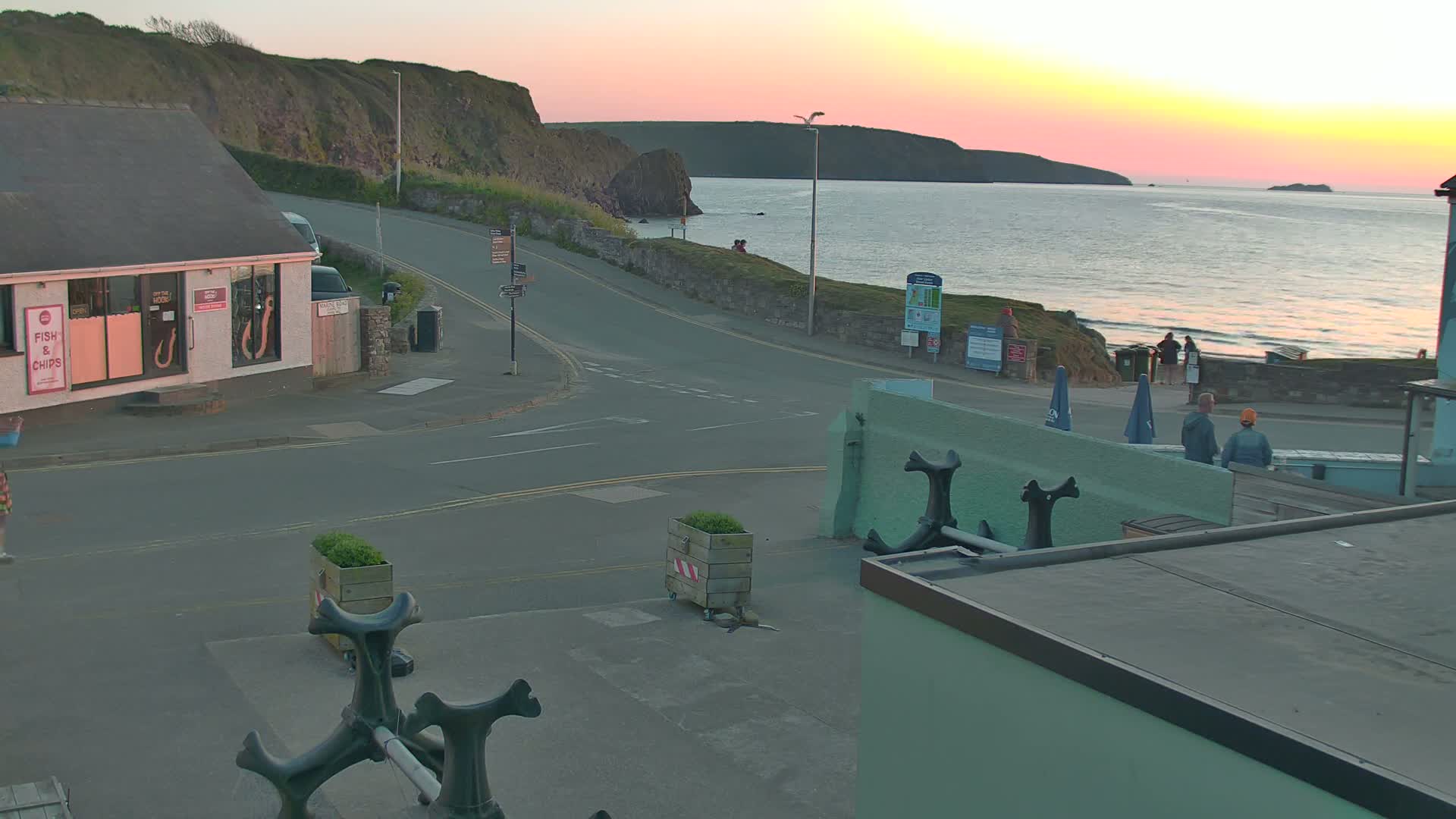 A coastal town scene at sunset shows a road curving toward a calm sea with a few people walking along the shore, near buildings and a fish and chips shop.