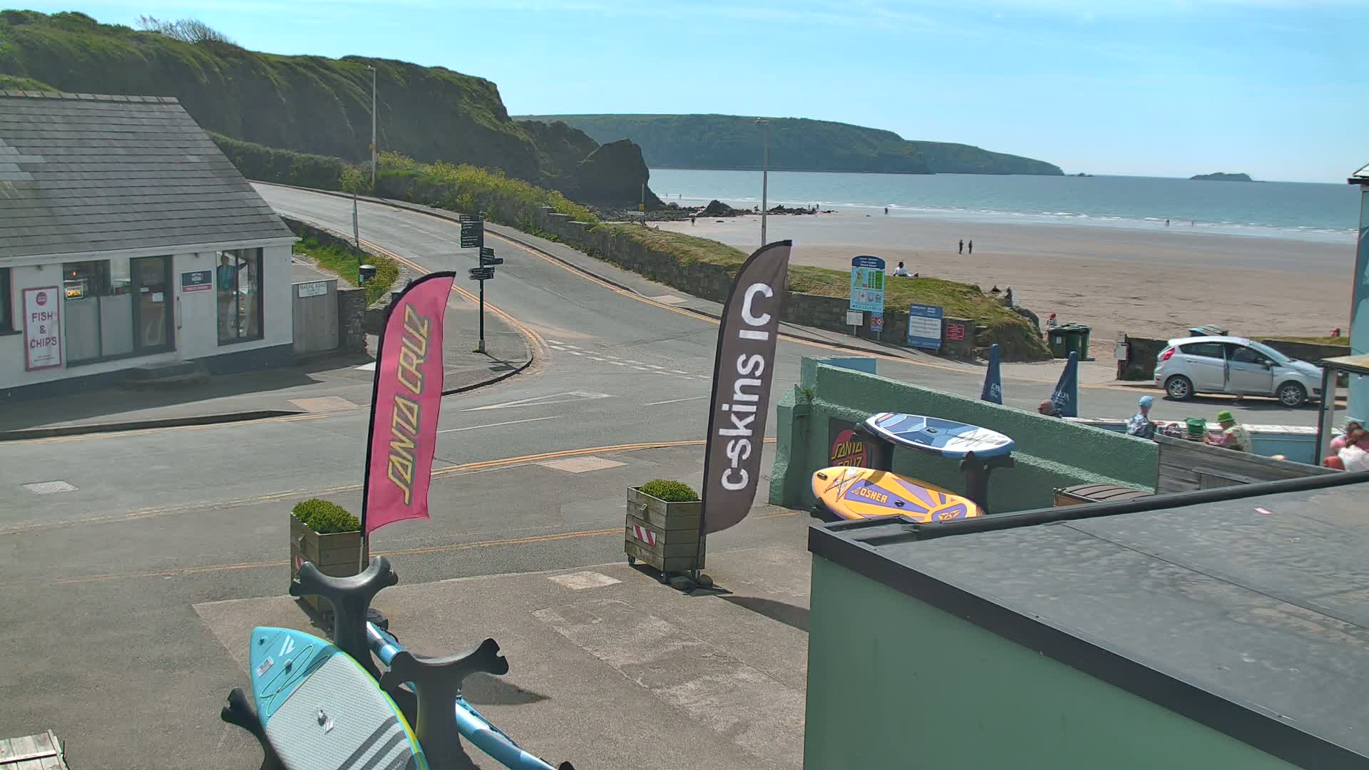 A sunny day shows a sandy beach with a few people, and a street scene with a fish and chips shop, some paddleboards, and a parked car.