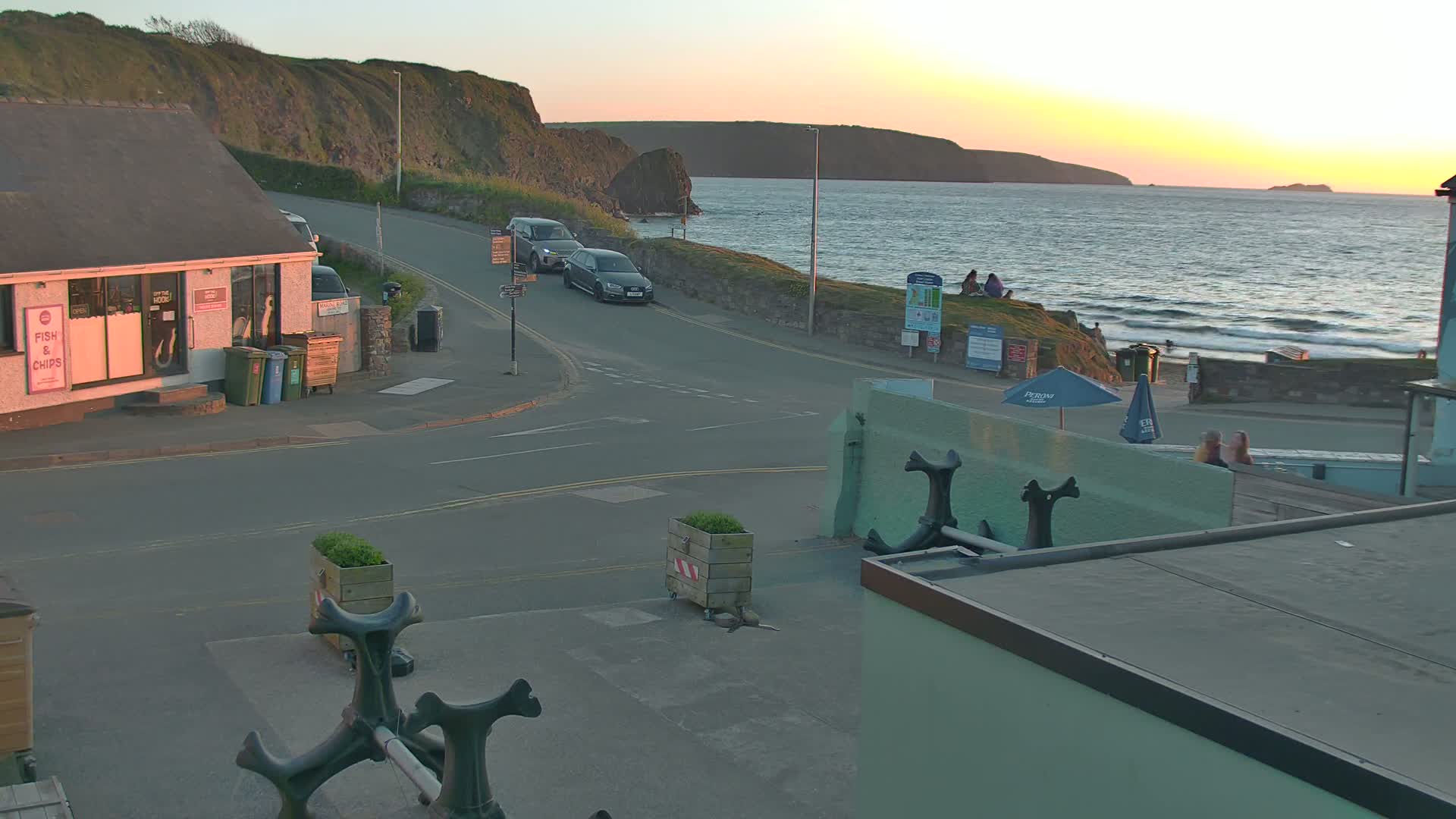 A seaside town at sunset, with cars parked near a fish and chips shop, overlooking a calm ocean with people sitting on a grassy area.