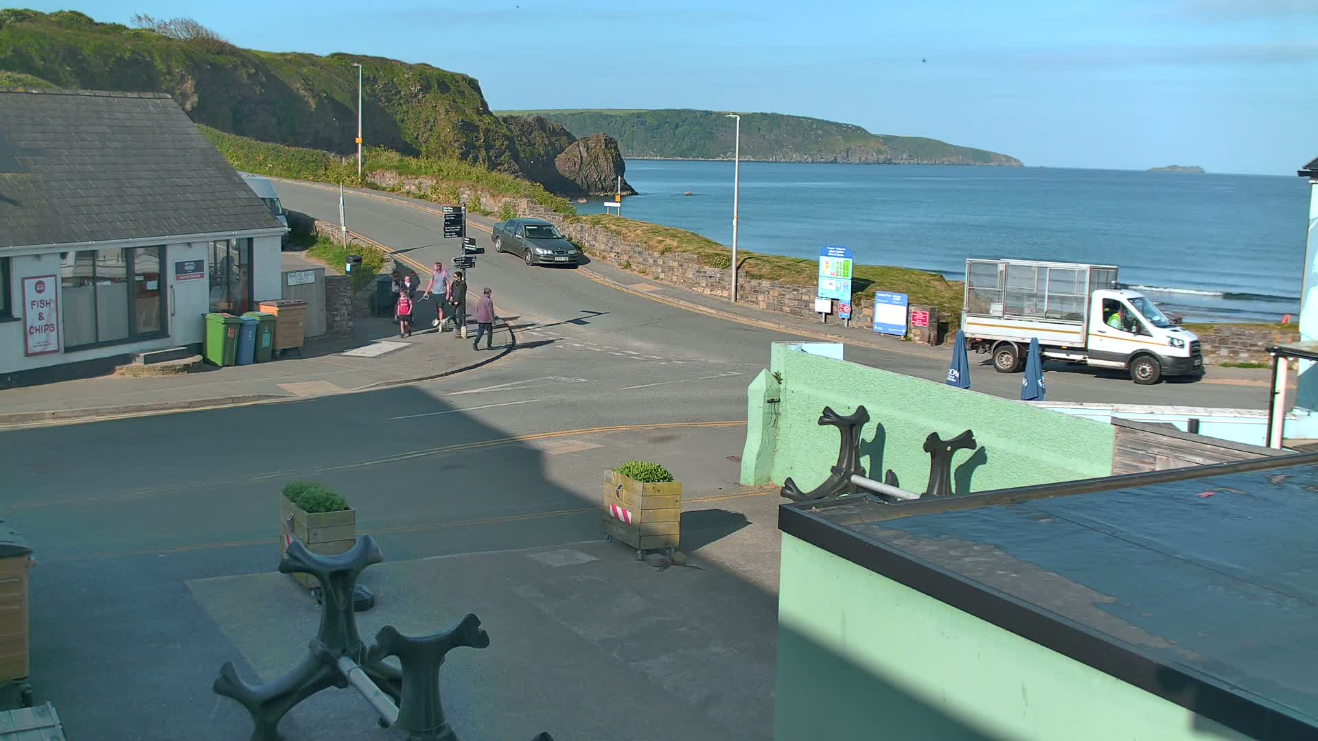 A sunny coastal road scene shows a few pedestrians near a fish and chips shop, a car, and a delivery truck parked near a beach with calm waves, all situated against a backdrop of cliffs and a sea.